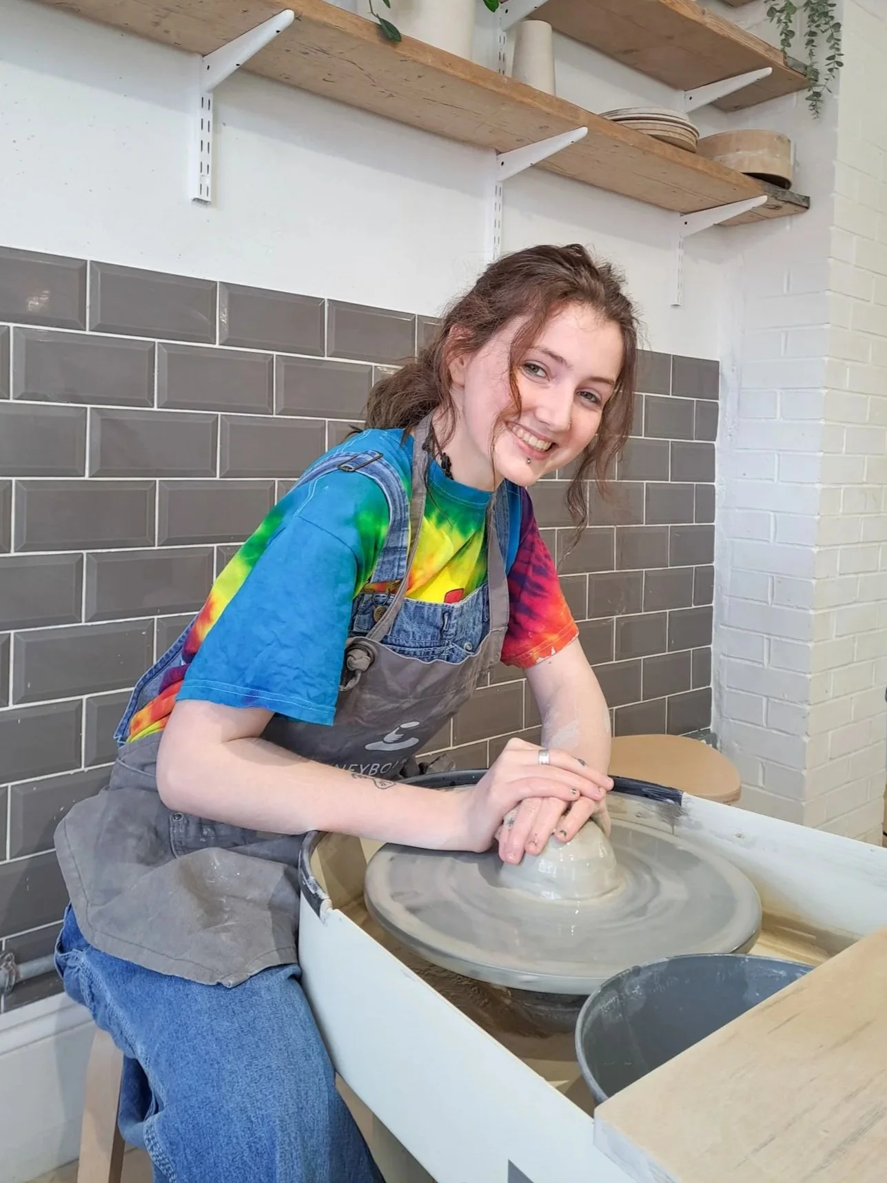 A young woman with a tie-dye shirt and apron, smiling while working on a pottery wheel in a ceramics studio.