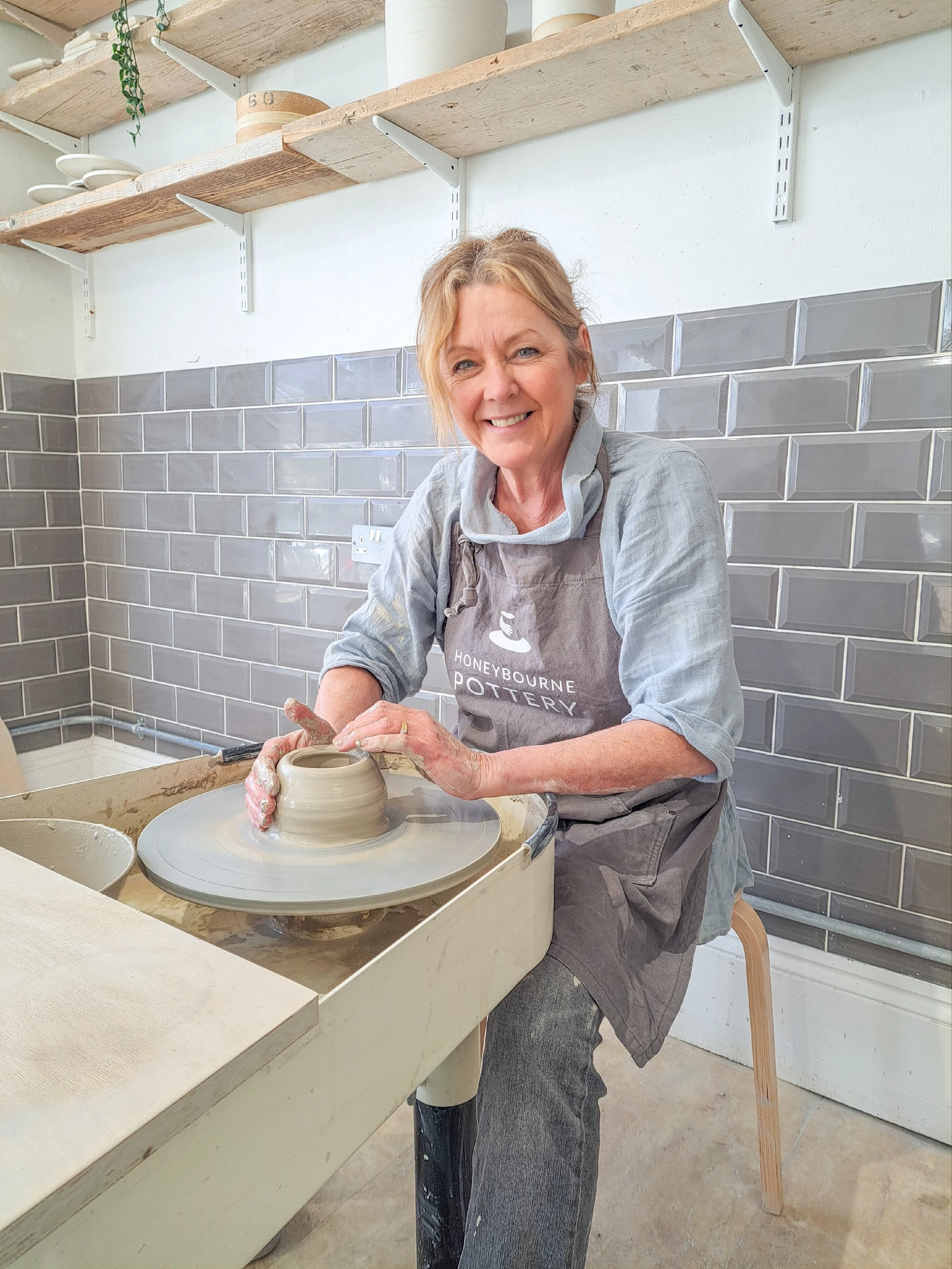 A smiling woman working on pottery at a wheel in a pottery studio, wearing a gray apron and a light gray shirt with shelves of white plates and bowls behind her.