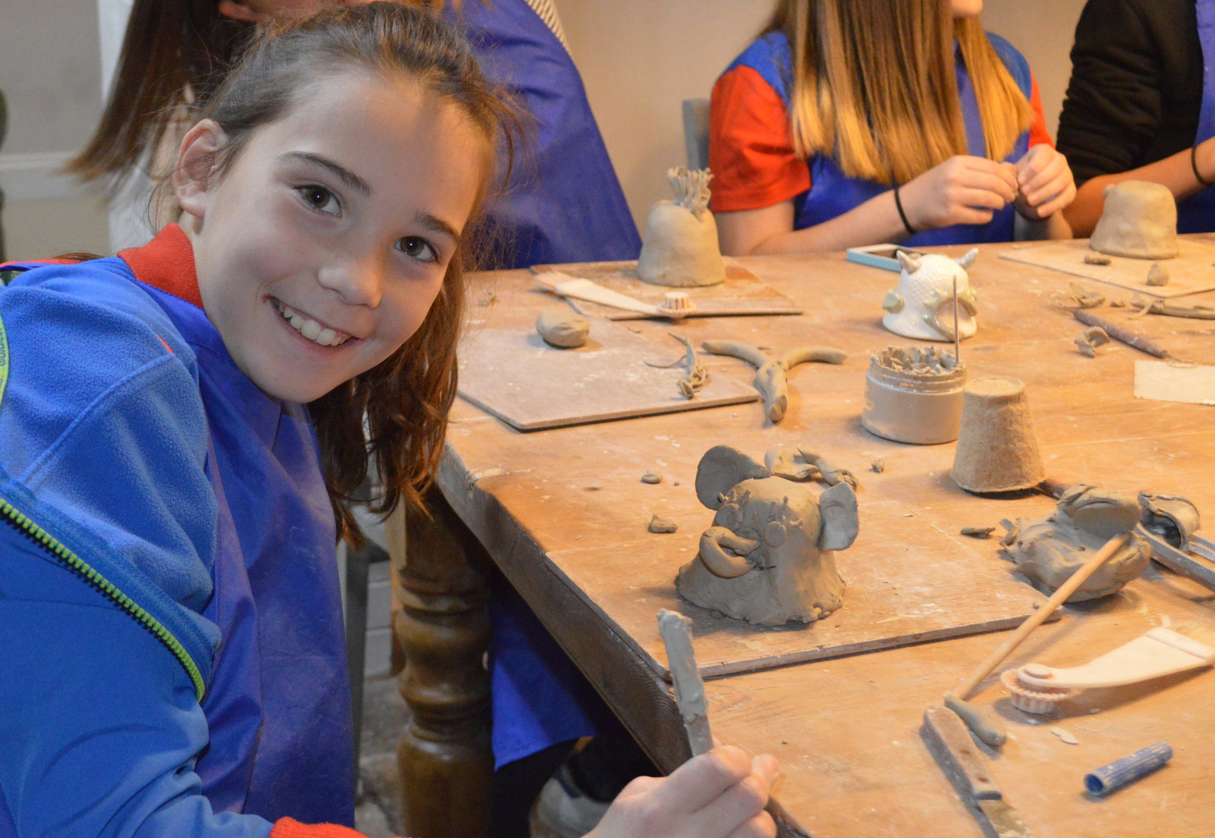 A young girl smiling and working on a clay sculpture at a table, with other children in the background also engaged in pottery activities.