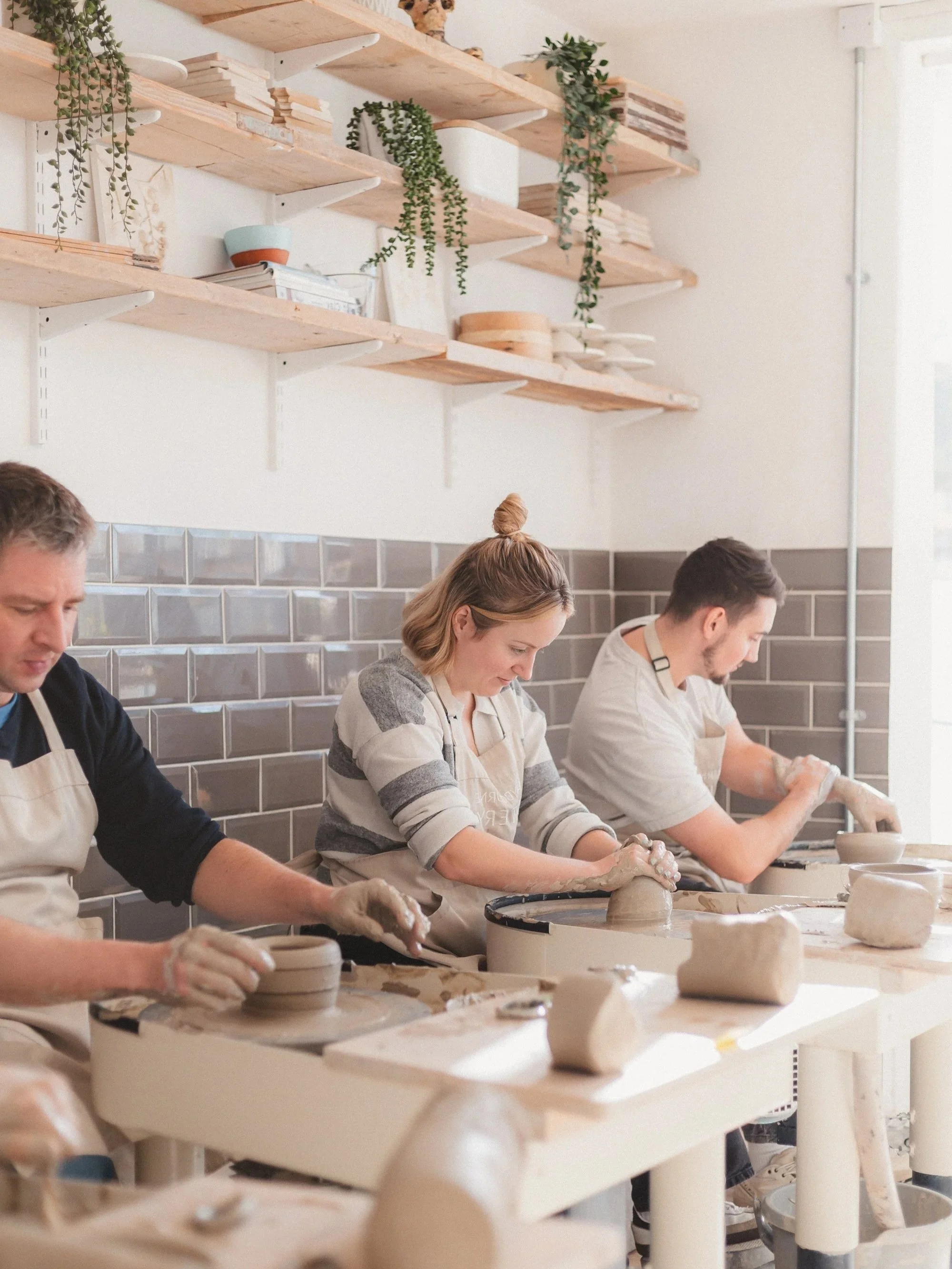 People participating in a pottery class, shaping clay on pottery wheels in a well-lit studio with wooden shelves and potted plants in the background.