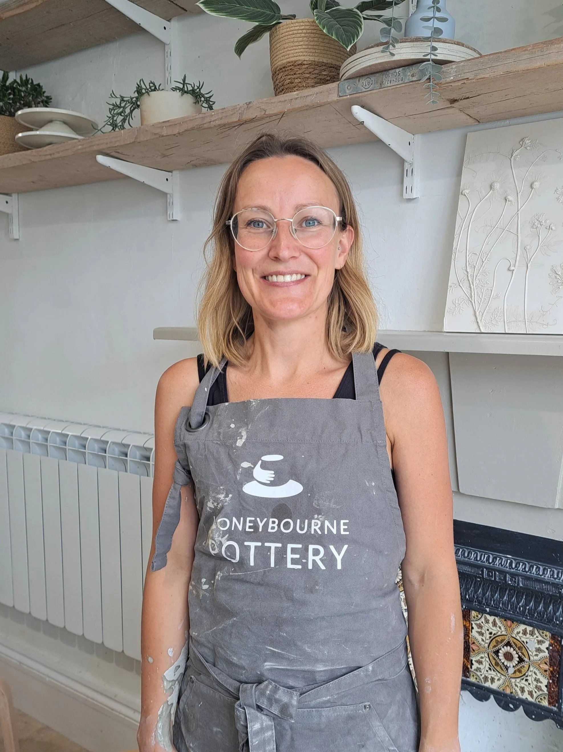 A woman with shoulder-length blonde hair and glasses smiling at the camera, wearing a gray apron with the words "Honeybourne Pottery" on it, standing in a pottery studio with shelves of pottery and plants in the background.