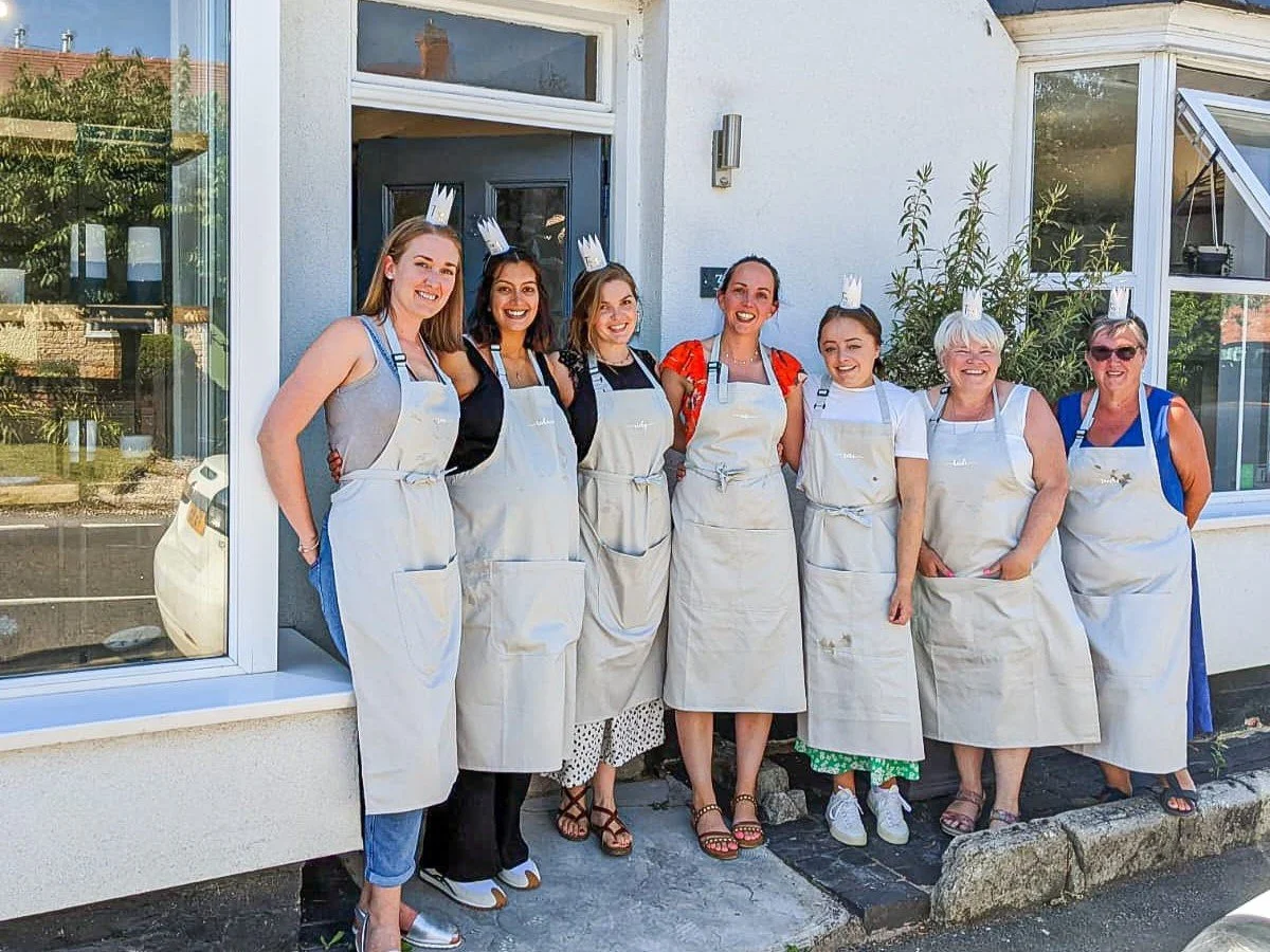 Group of seven women standing outside Honeybourne Pottery in aprons and paper crowns, smiling for the photo.
