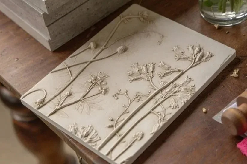 A clay or plaster bas-relief artwork of pressed and carved botanical plants and flowers on a square tile, resting on a wooden table.