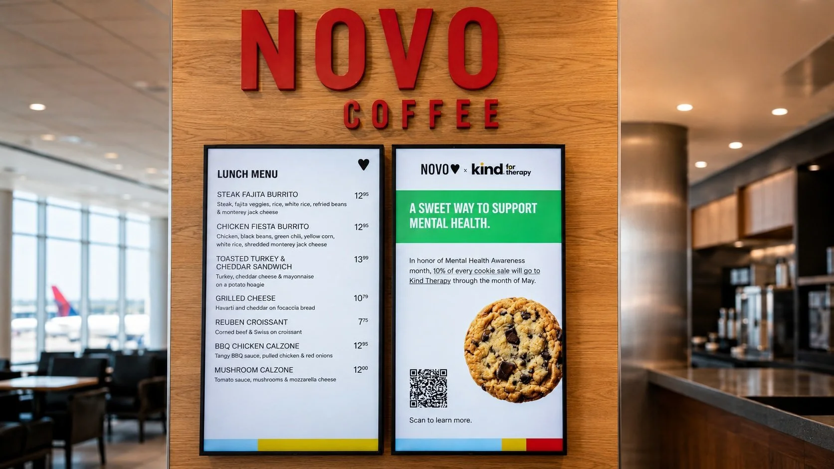 Digital menu screens inside a Novo Coffee location. One screen shows the lunch menu, while the other promotes a Novo Coffee × Kind Therapy Inc campaign reading “A sweet way to support mental health,” with a chocolate chip cookie image and a QR code.