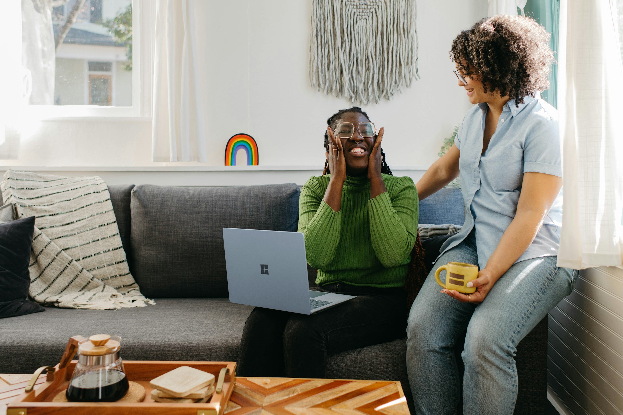 Two people smile on a gray couch during a laptop video call, one reacting with excitement while the other holds a yellow mug in a bright living room with a rainbow decoration.