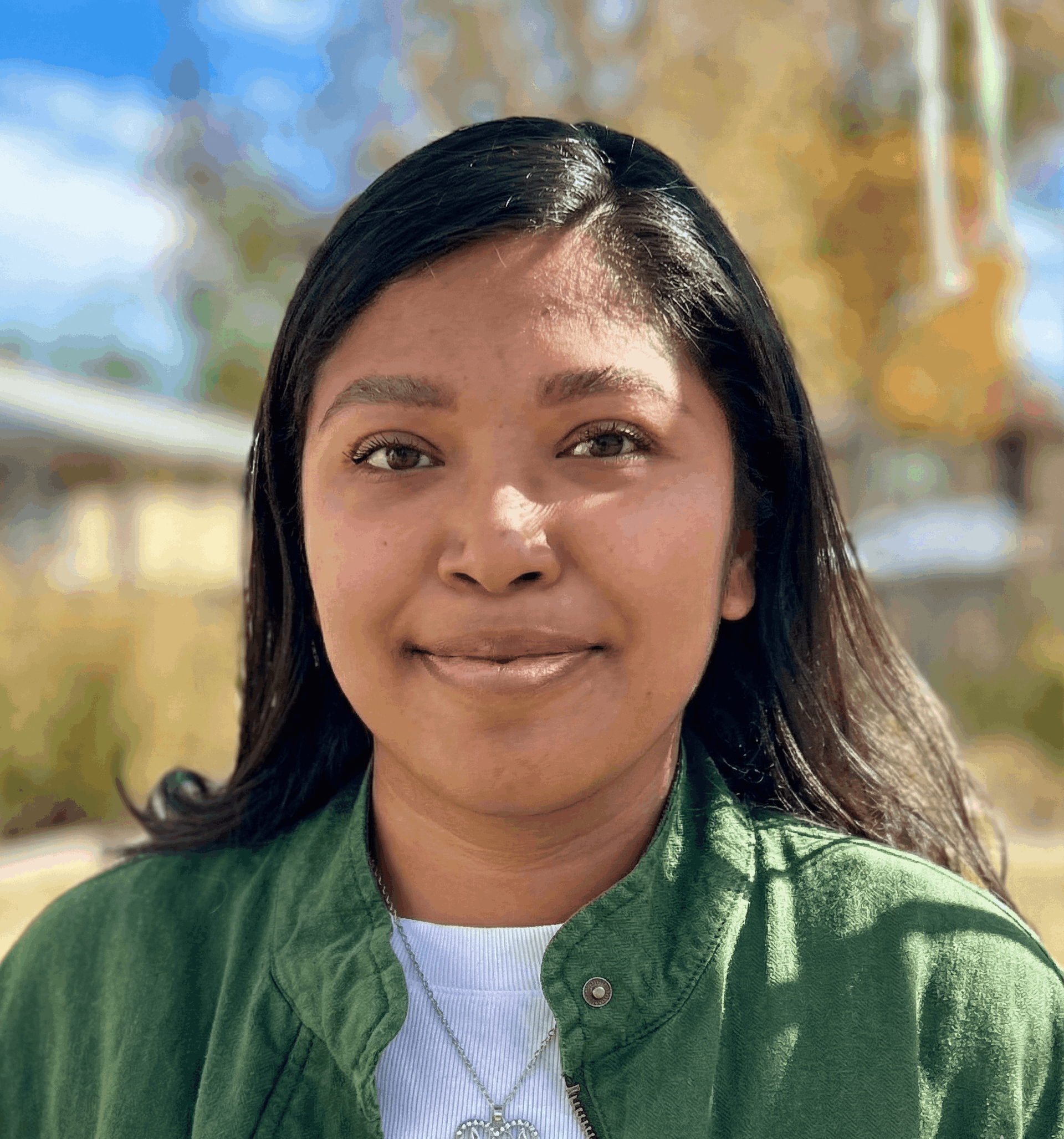 person with long dark hair, green collared shirt and a necklace smiling, standing outdoors