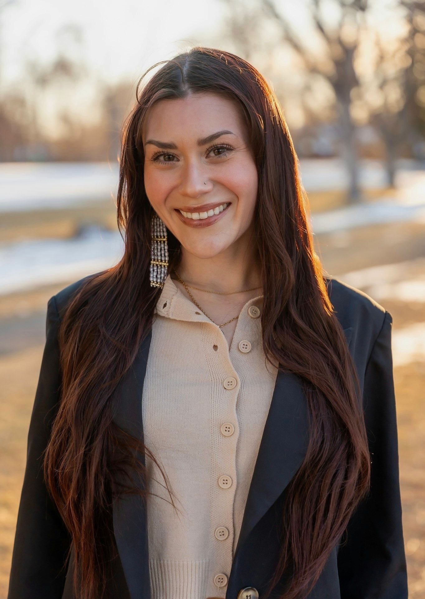 Person with long dark hair smiling, wearing a bright blue dress, standing outdoors in front of flowering trees and a fountain