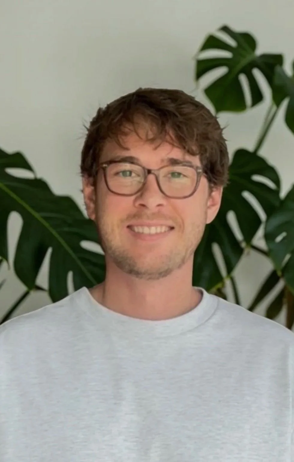 A smiling person wearing glasses and a light grey sweater, standing in front of a plant background.