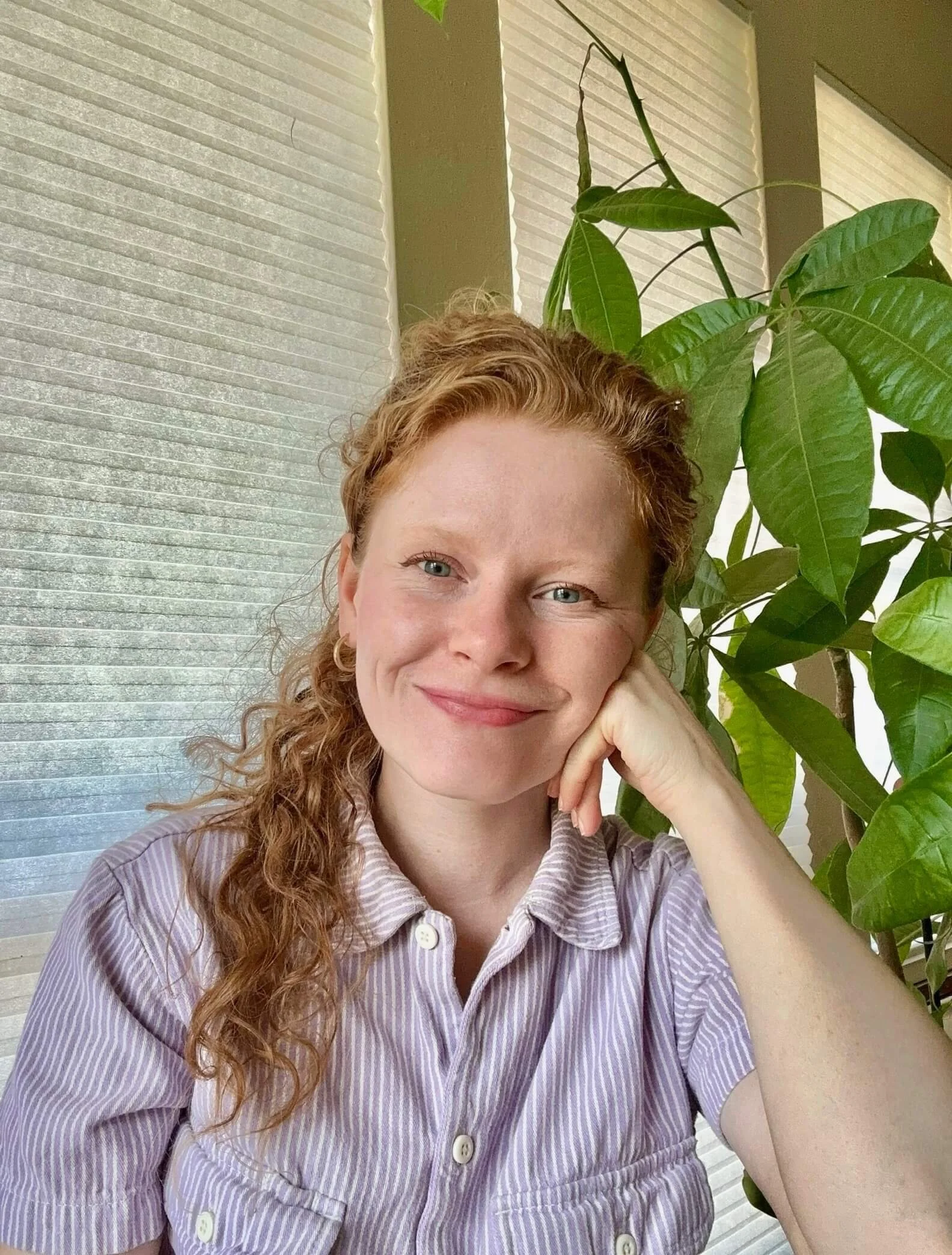 Person smiling in color photo, wearing a collared shirt with their head resting on their left hand in front of a window and a plant