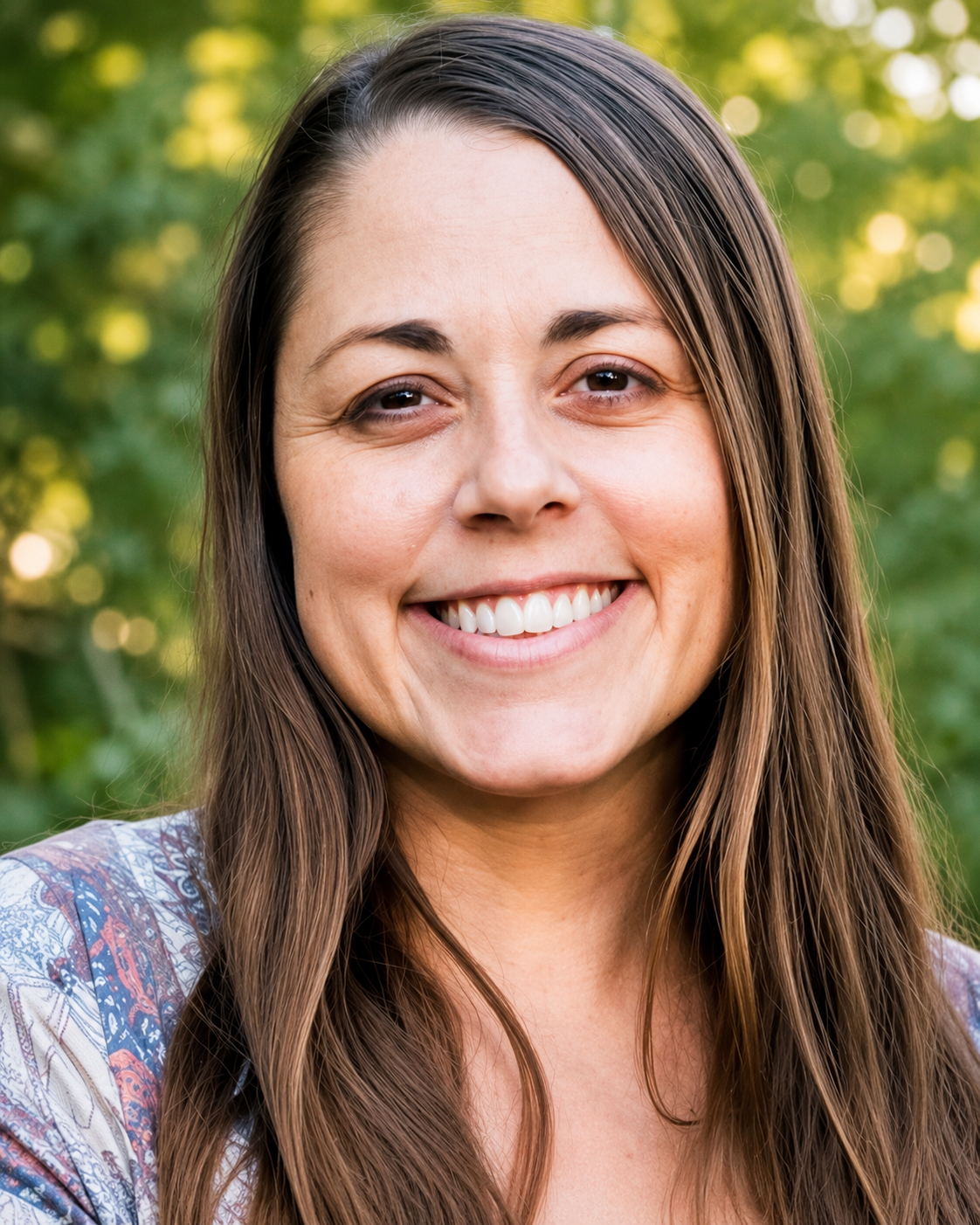 Close-up outdoor headshot of a smiling woman with long brown hair wearing a patterned top, photographed against a softly blurred green nature background with warm natural lighting