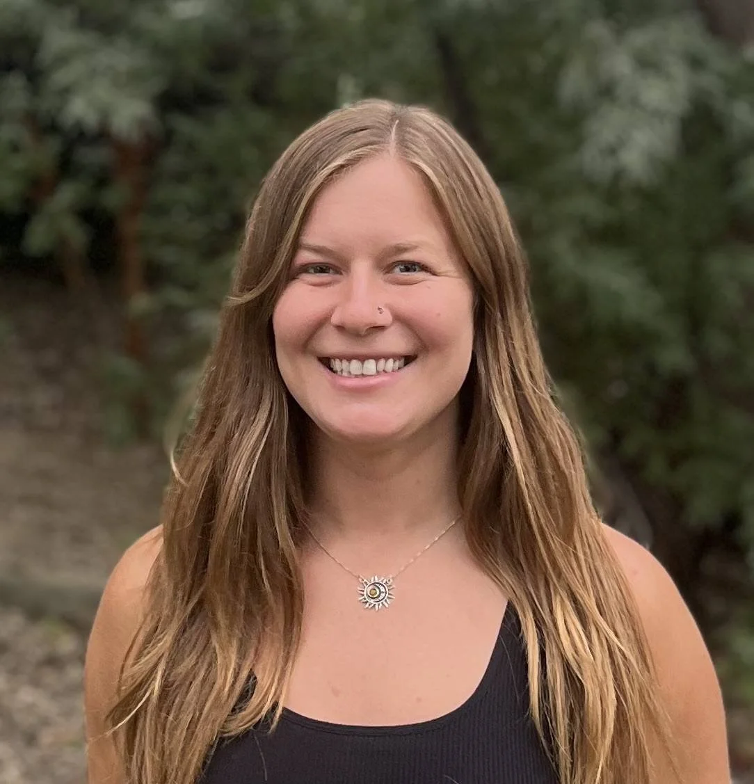 person with long dark blonde hair over their shoulders smiling standing in front of trees and rocks