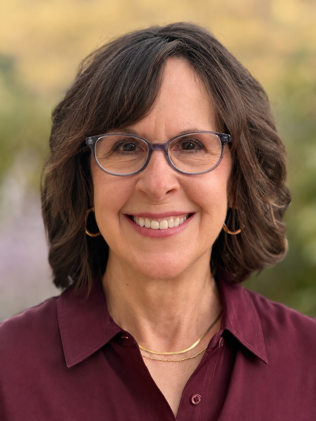 Person with shoulder-length brown hair and glasses smiling and wearing a maroon button-down shirt against a soft, blurred background.