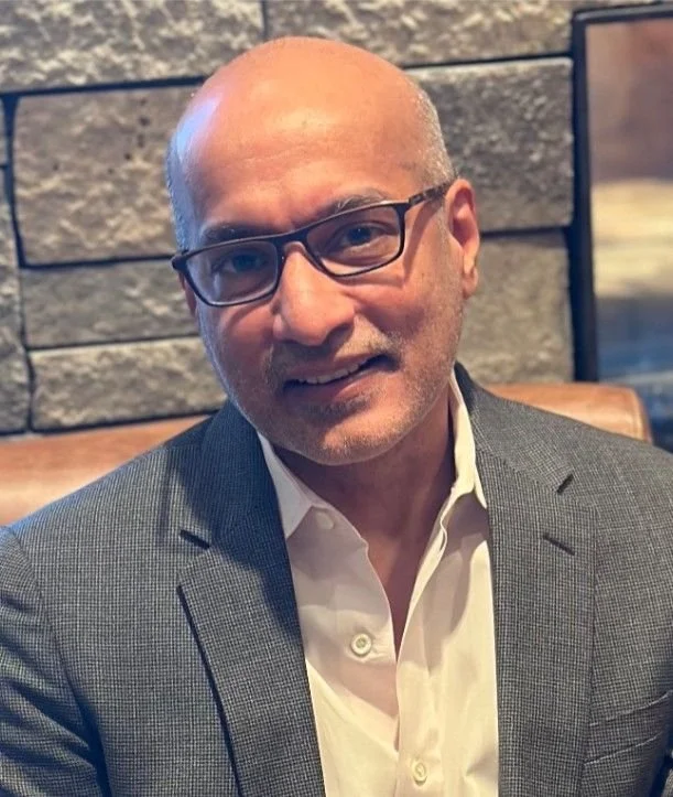 Person with no hair and glasses smiling in front of a grey brick wall wearing a grey suit with a white undershirt