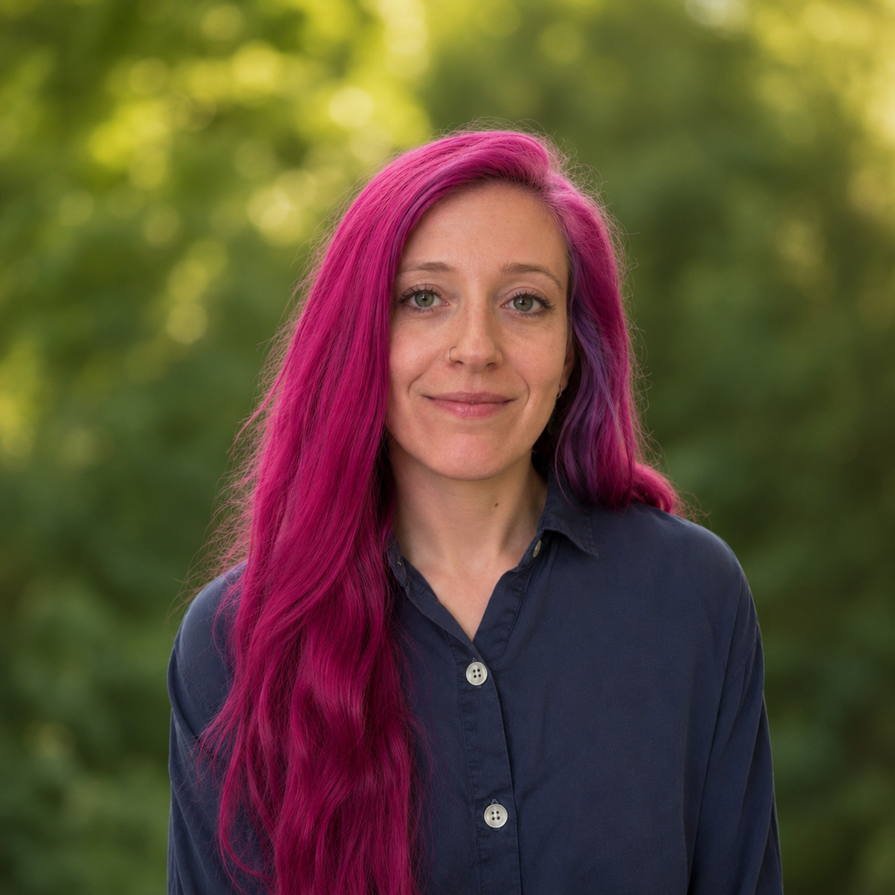 person with purple hair and blue button up shirt smiling in front of a green nature scene. 