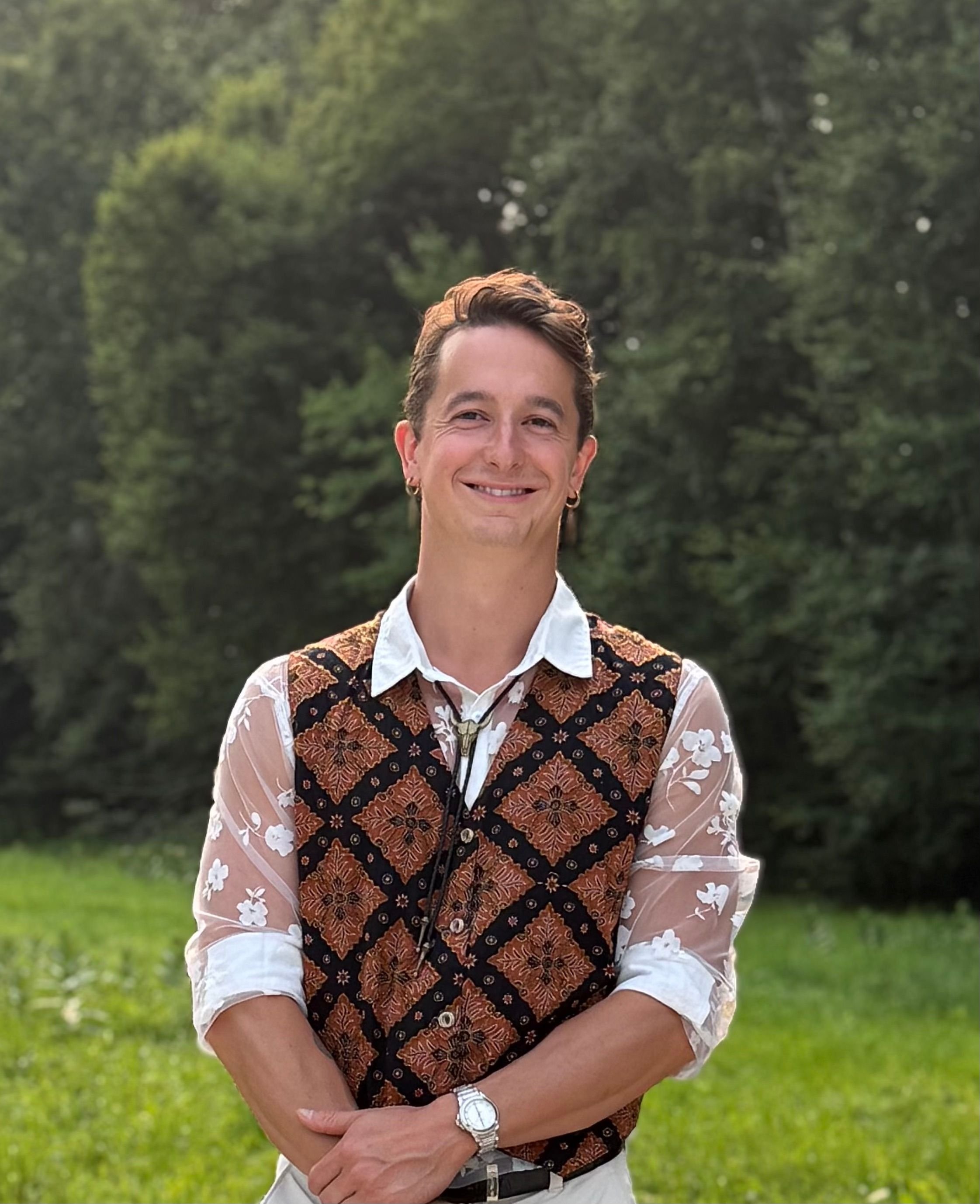 person smiling in color photo with short brown hair, in a vest with rolled up sleeves, arms crossed, wearing a watch