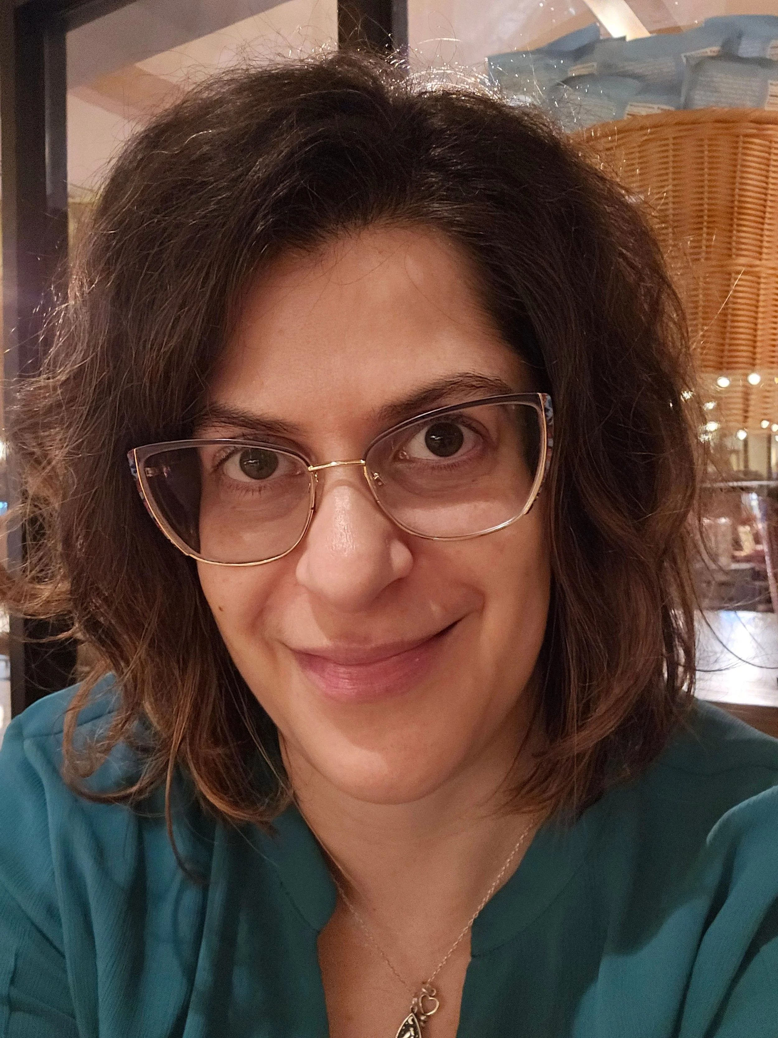 Person with shoulder length brown hair with glasses and a blue shirt with a necklace smiling. 