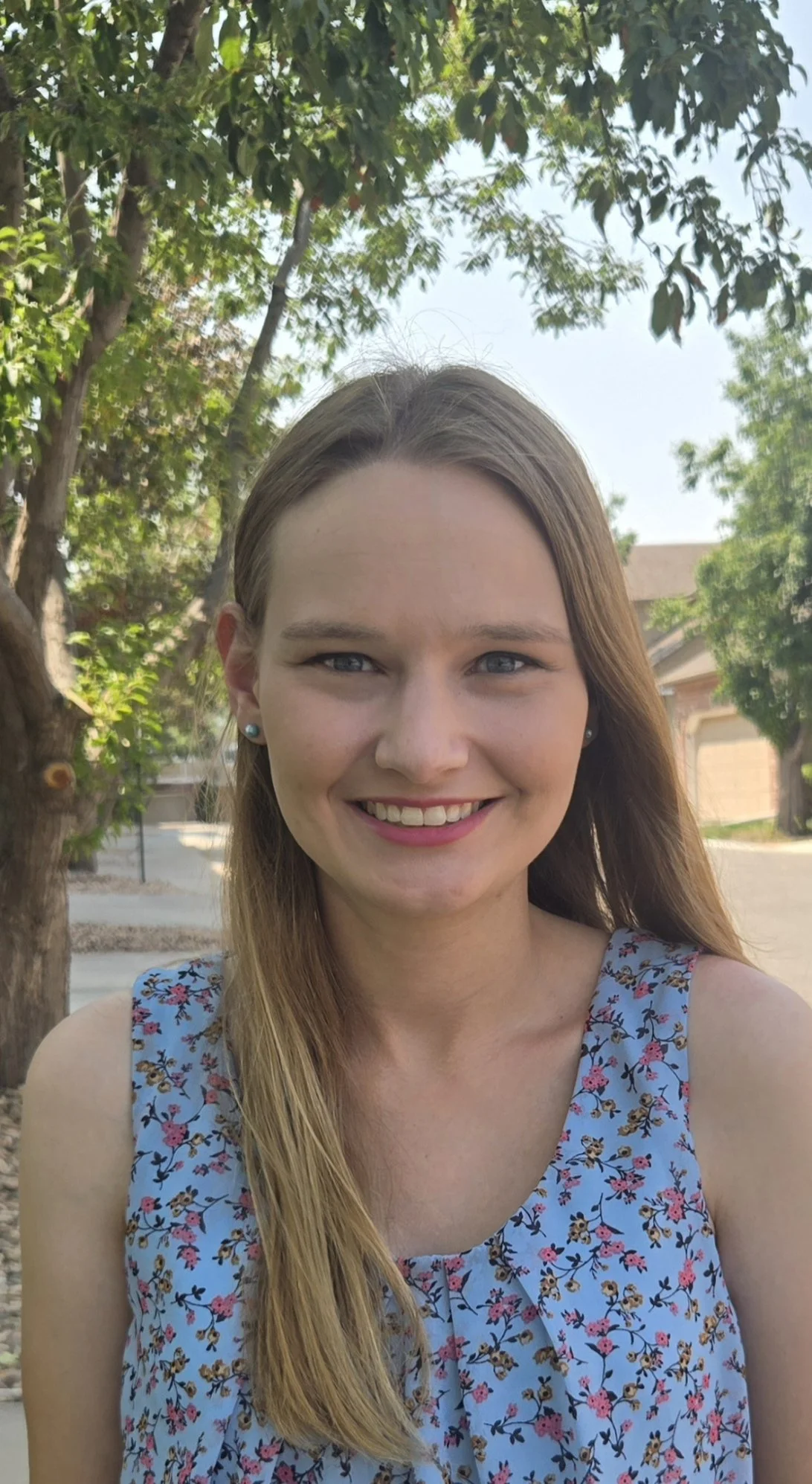 A smiling young person with long blonde hair, wearing a light blue floral top, standing outdoors in front of trees.