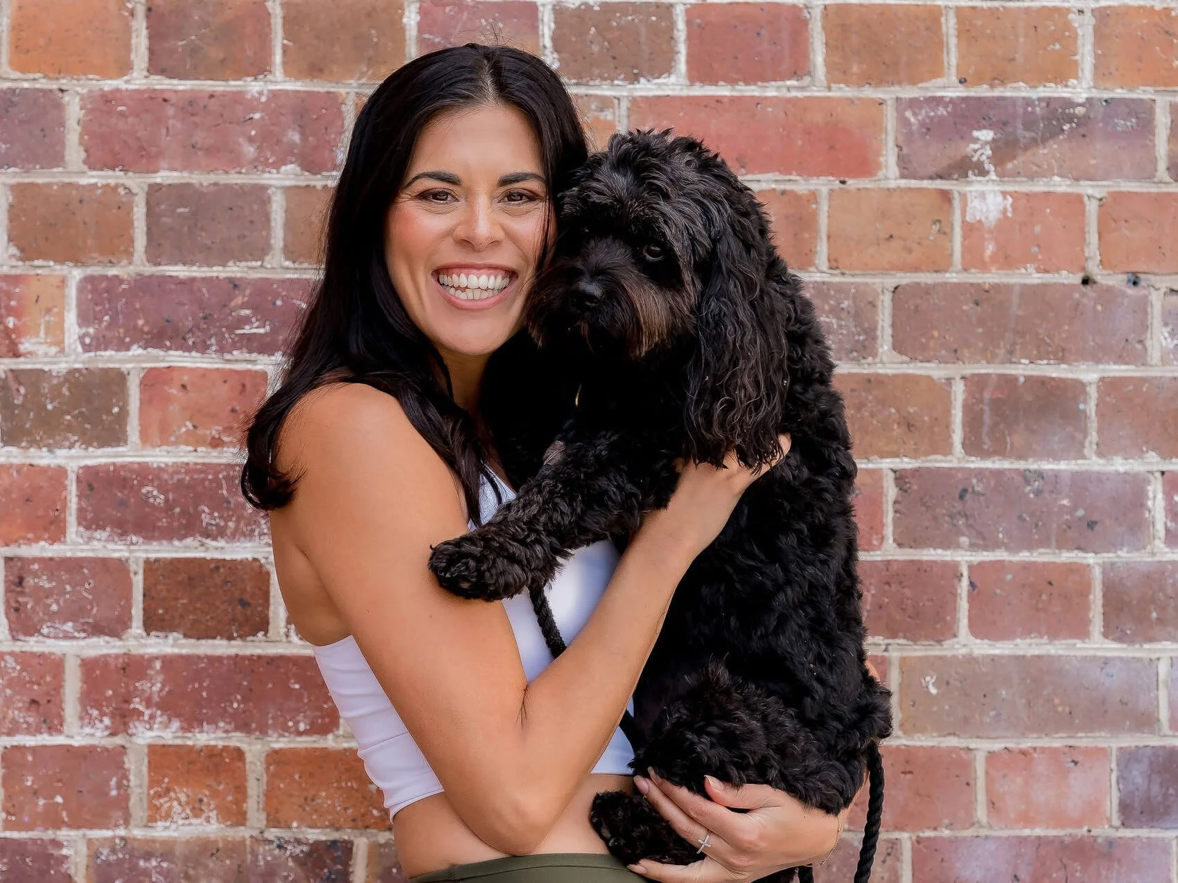 A woman with dark hair smiling and holding a black, curly-haired dog in front of a brick wall.