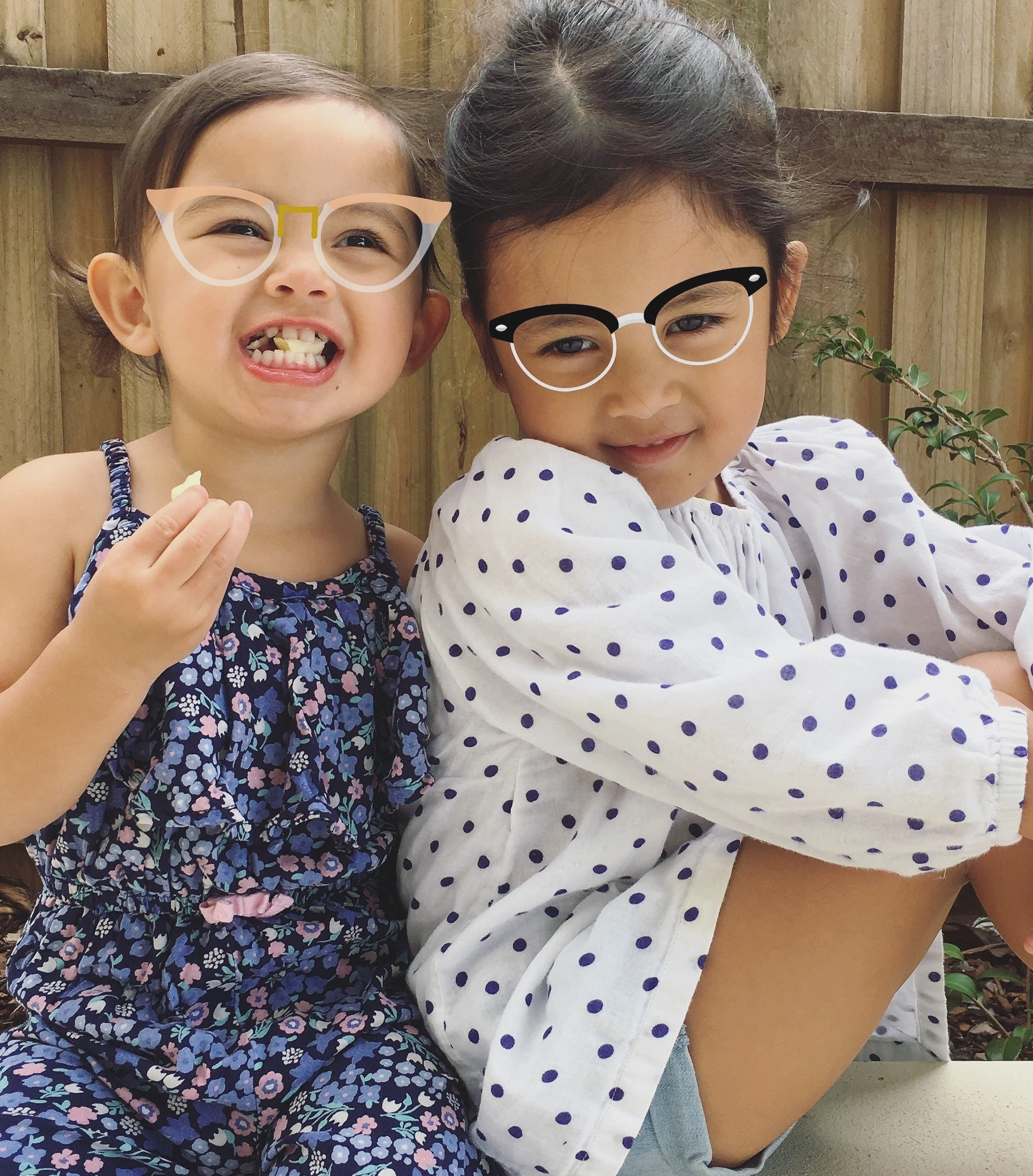 Two young girls sitting outdoors, wearing glasses with playful photo filter effects, smiling at the camera. One girl is holding a snack.