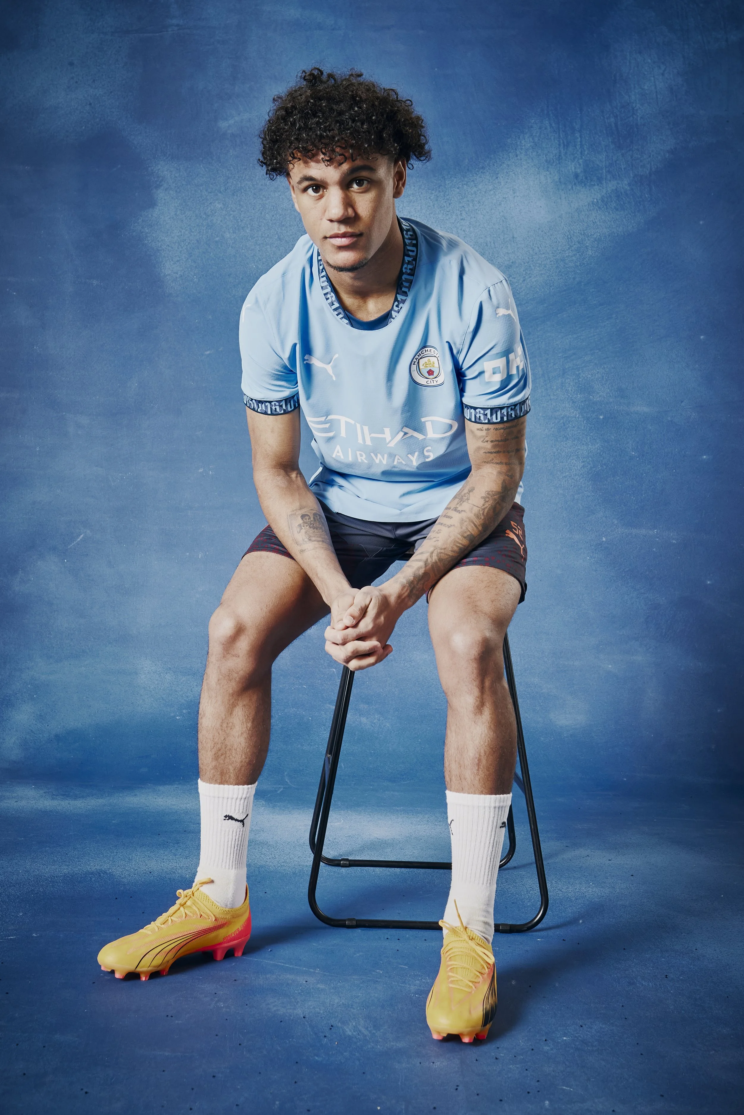 A young man with curly hair is sitting on a chair against a blue background. He is wearing a light blue Manchester City soccer jersey, white Puma socks, and yellow soccer cleats.