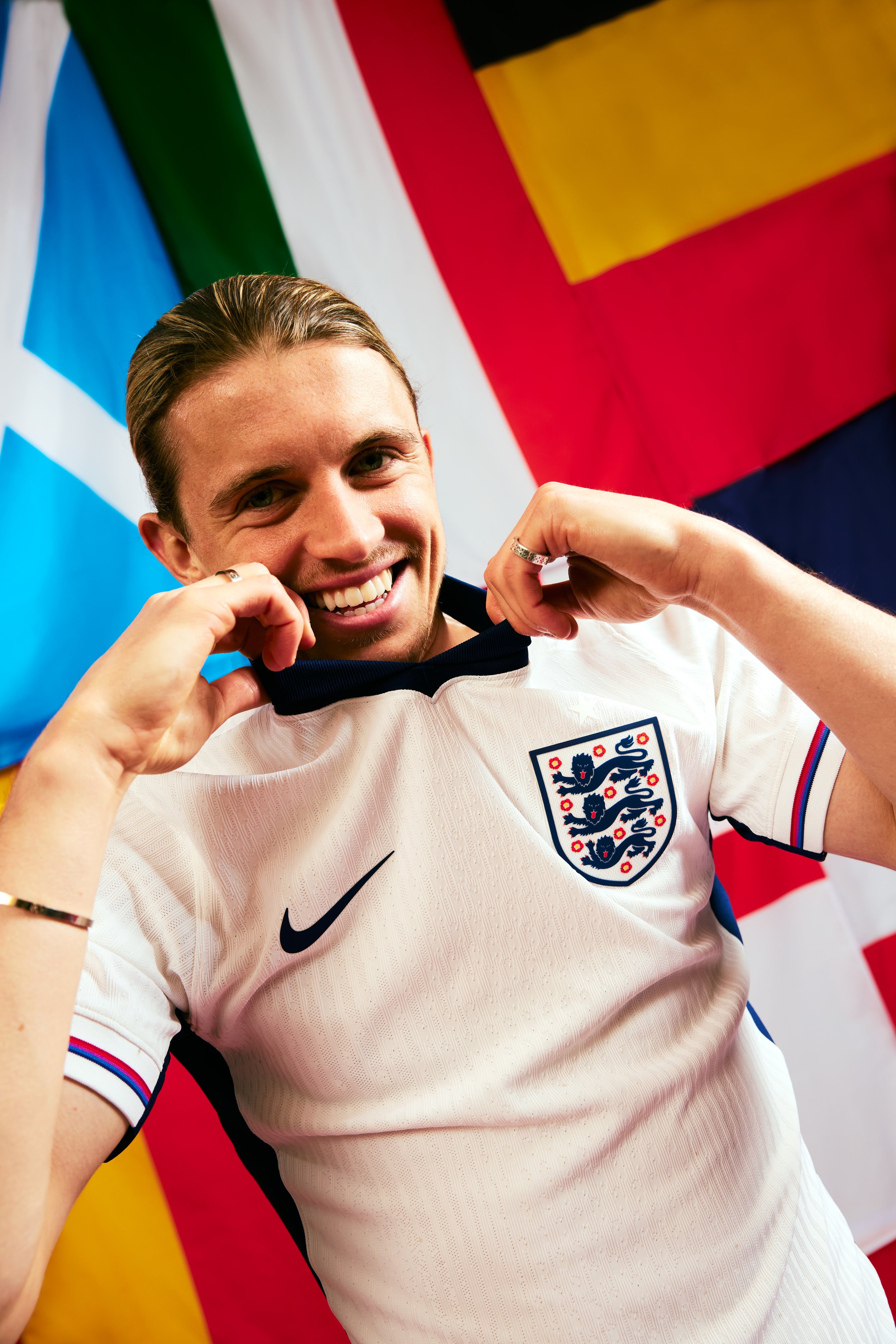 A smiling man in an England football jersey holding the collar of his shirt with flags of Greece, Italy, and another flag in the background.