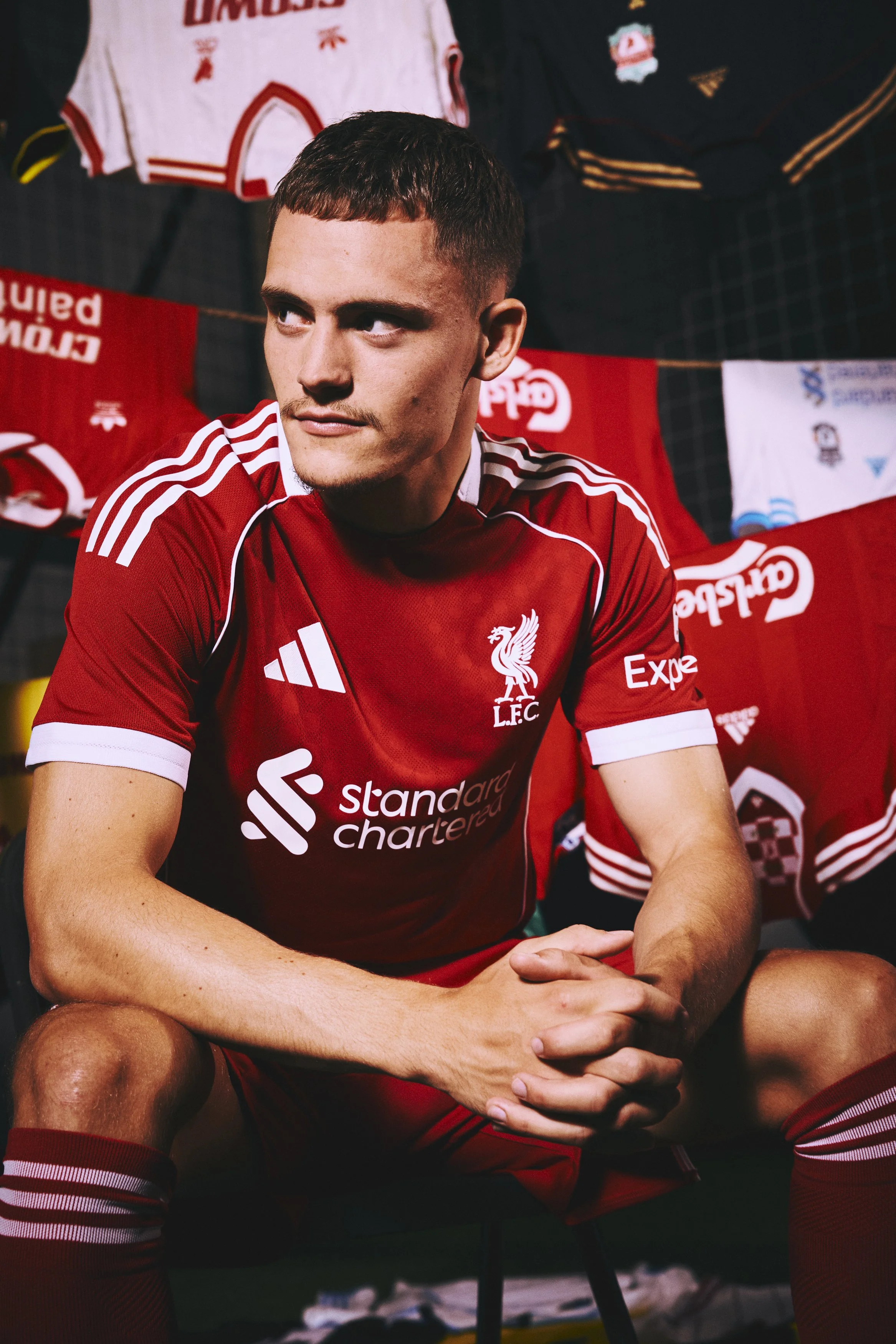 A man in a red Liverpool football jersey sitting with his hands clasped, surrounded by Liverpool jerseys and memorabilia.
