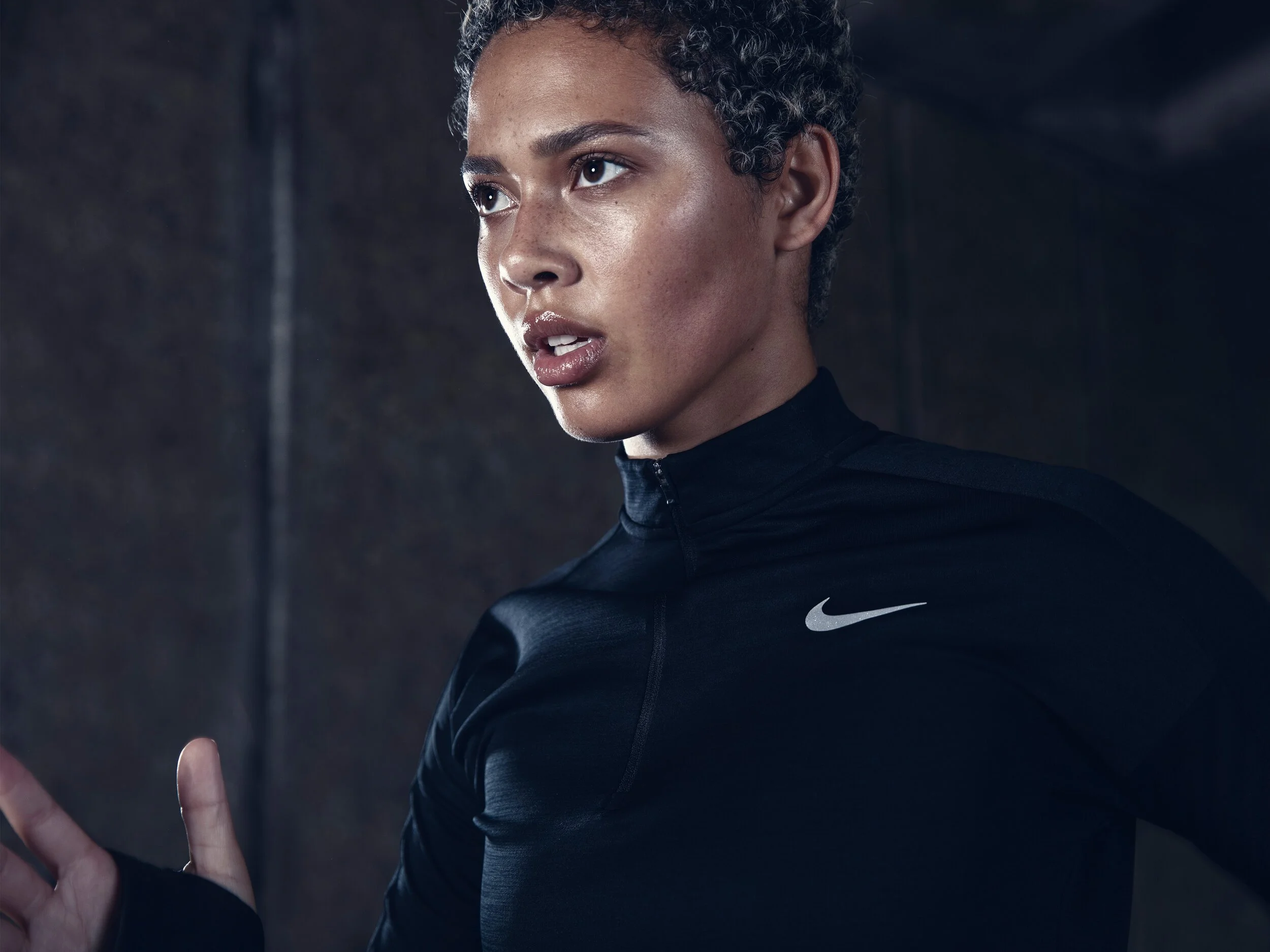 A woman wearing a black Nike sports shirt with a high collar, looking intently to her left, with a dark background.