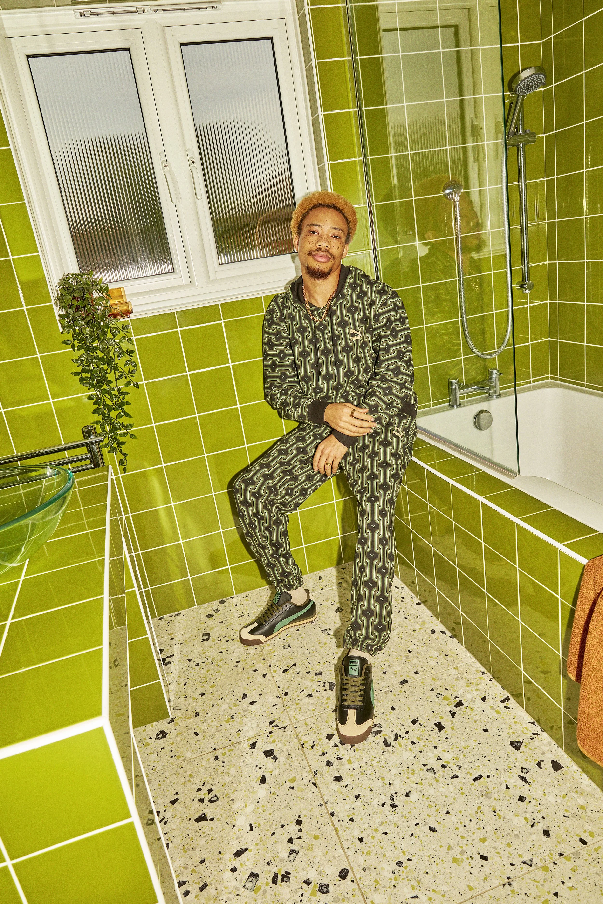 A young man with blond hair and face tattoos sitting on the edge of a bathtub in a green-tiled bathroom, wearing a black and green patterned tracksuit and sneakers.