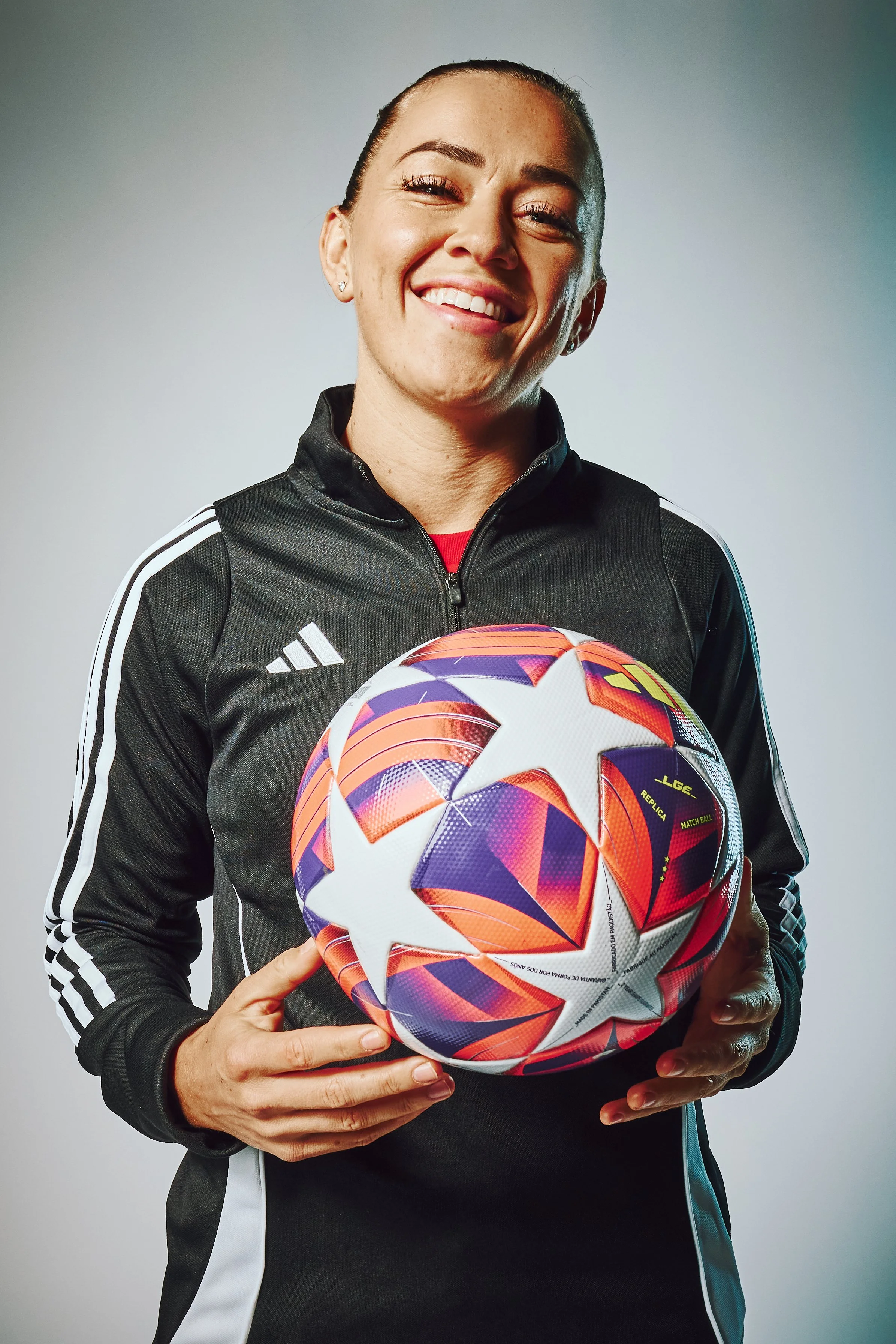A woman in a black Adidas soccer jacket holding a colorful soccer ball with stars and vibrant patterns, smiling at the camera.