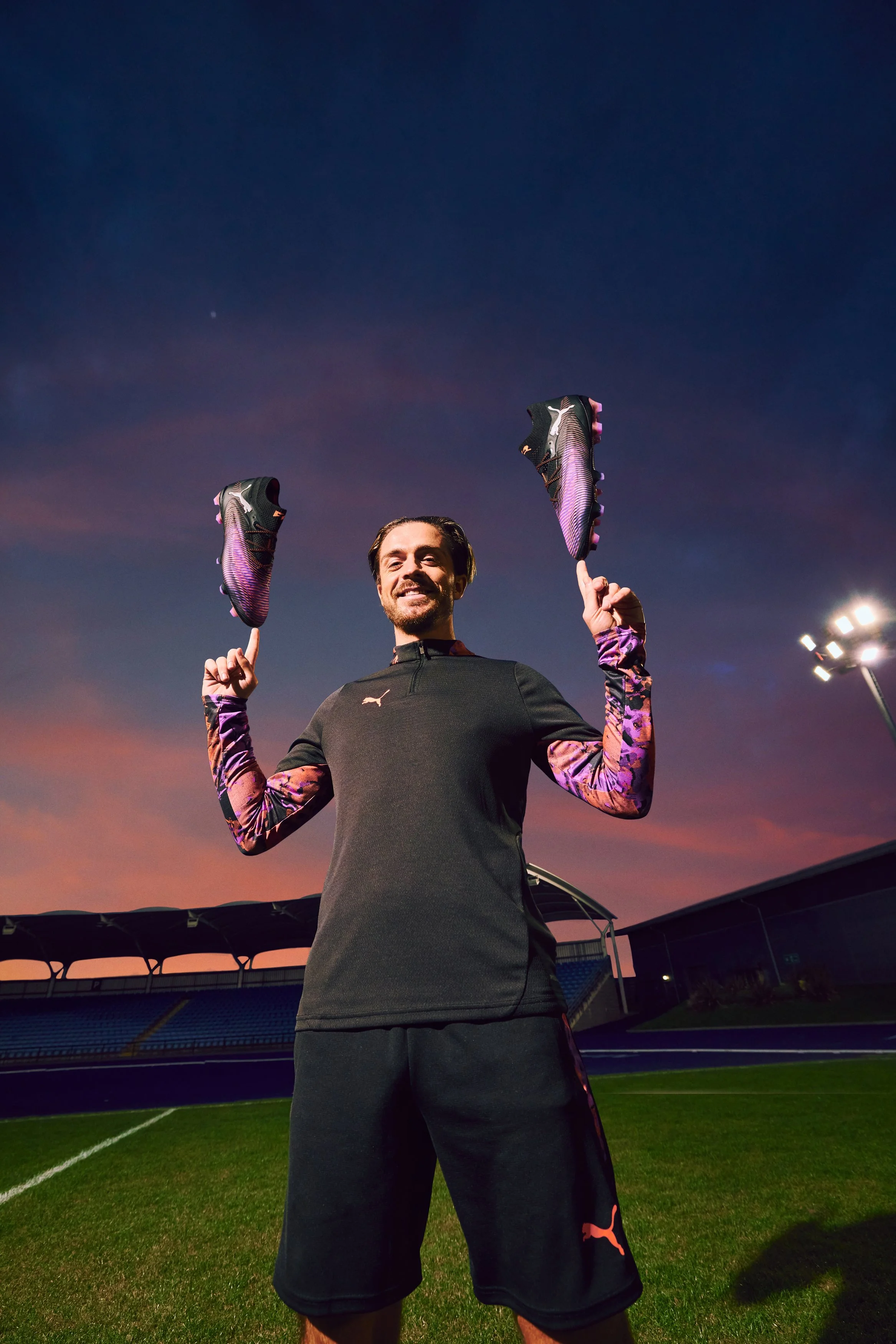 A man on a sports field during sunset, holding up a pair of purple soccer cleats and smiling.