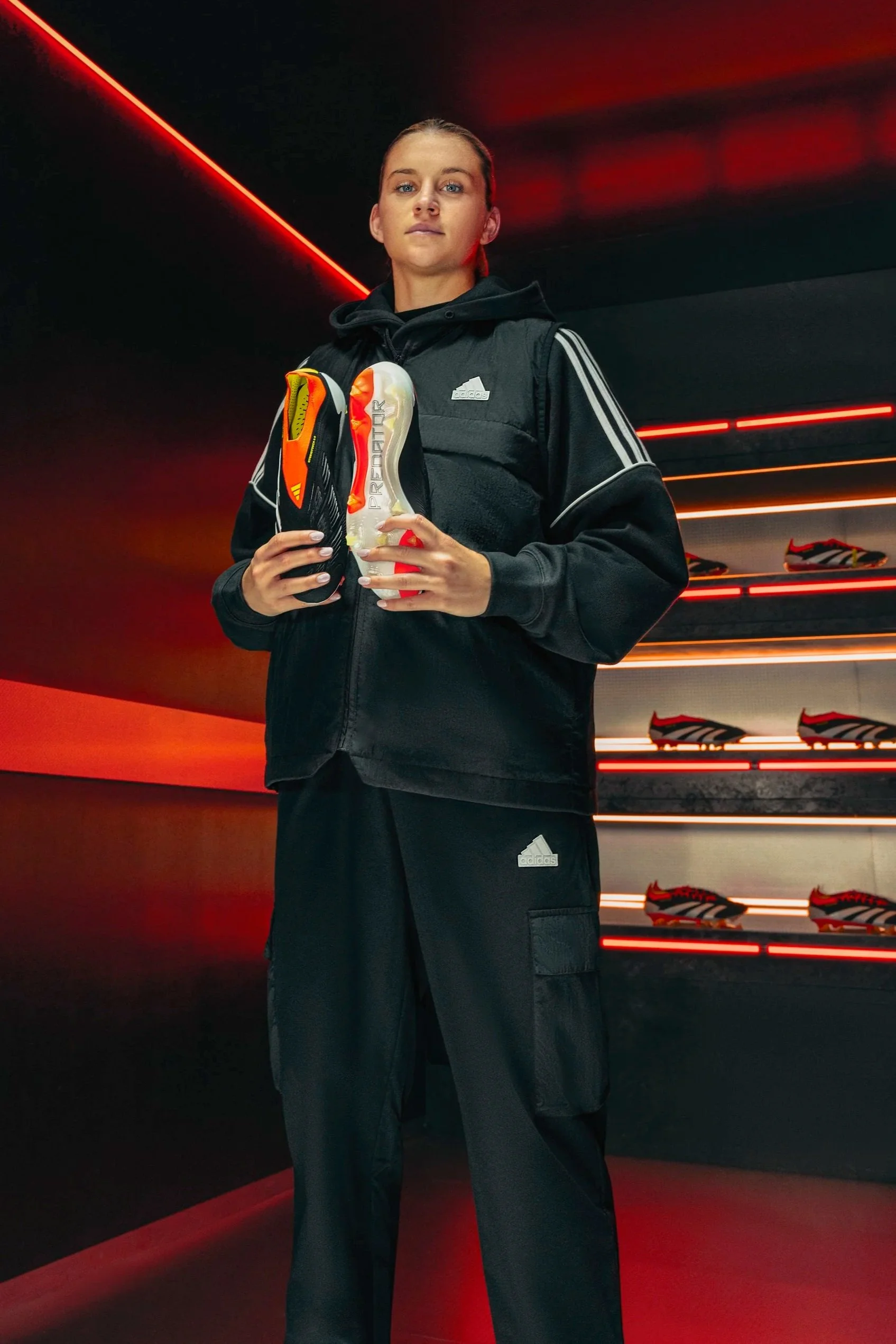A woman dressed in black Adidas athletic wear stands in a sports store holding a pair of brightly colored athletic shoes, with a background of soccer cleats on illuminated shelves.