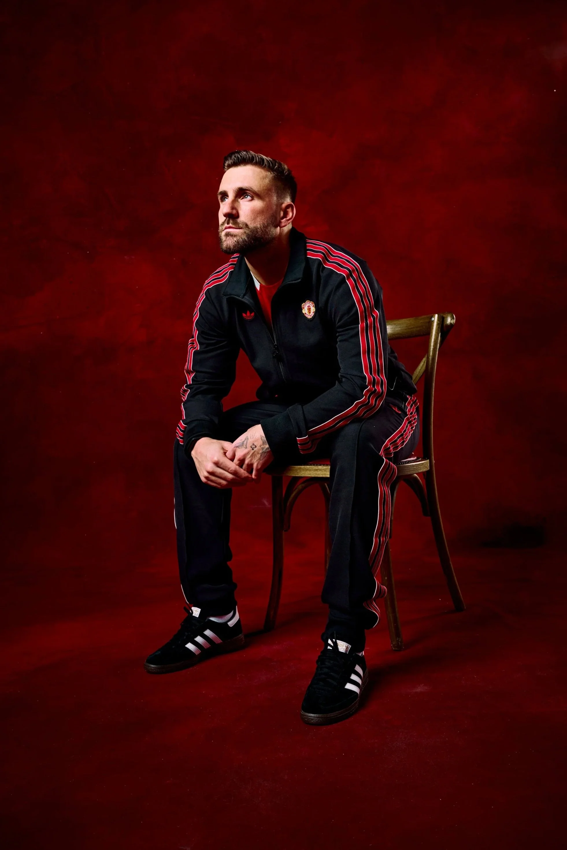 A man with a beard and short hair, wearing a black Adidas tracksuit with red stripes and a Manchester United logo, sitting on a wooden chair against a red background.