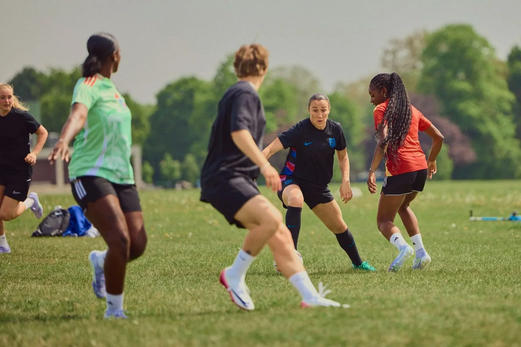 Women playing soccer on a grassy field during daytime with trees in the background, some players wearing athletic gear and shorts.