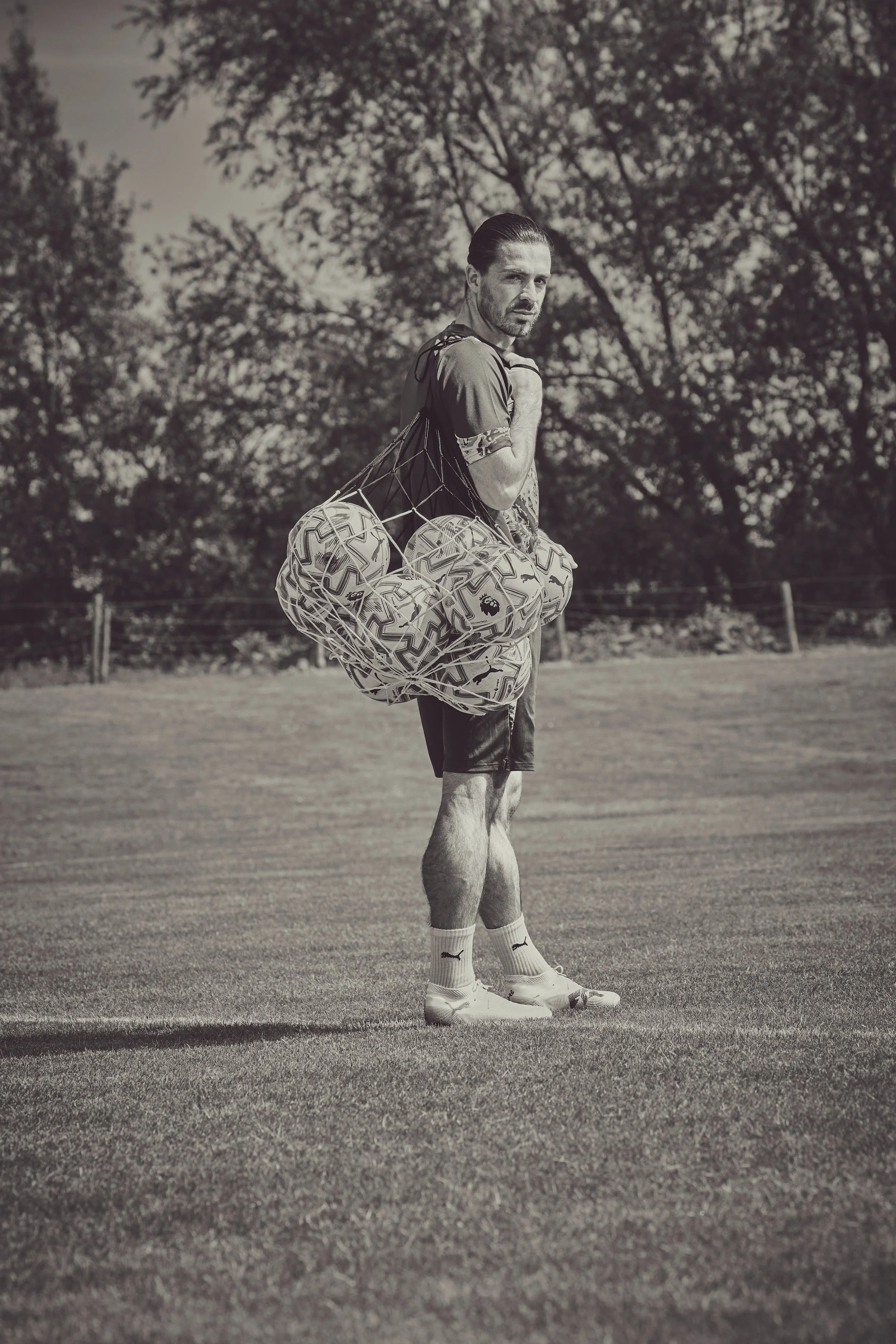 A man standing on a grassy field carrying a large bag filled with soccer balls, wearing athletic clothing and shoes, with trees in the background.