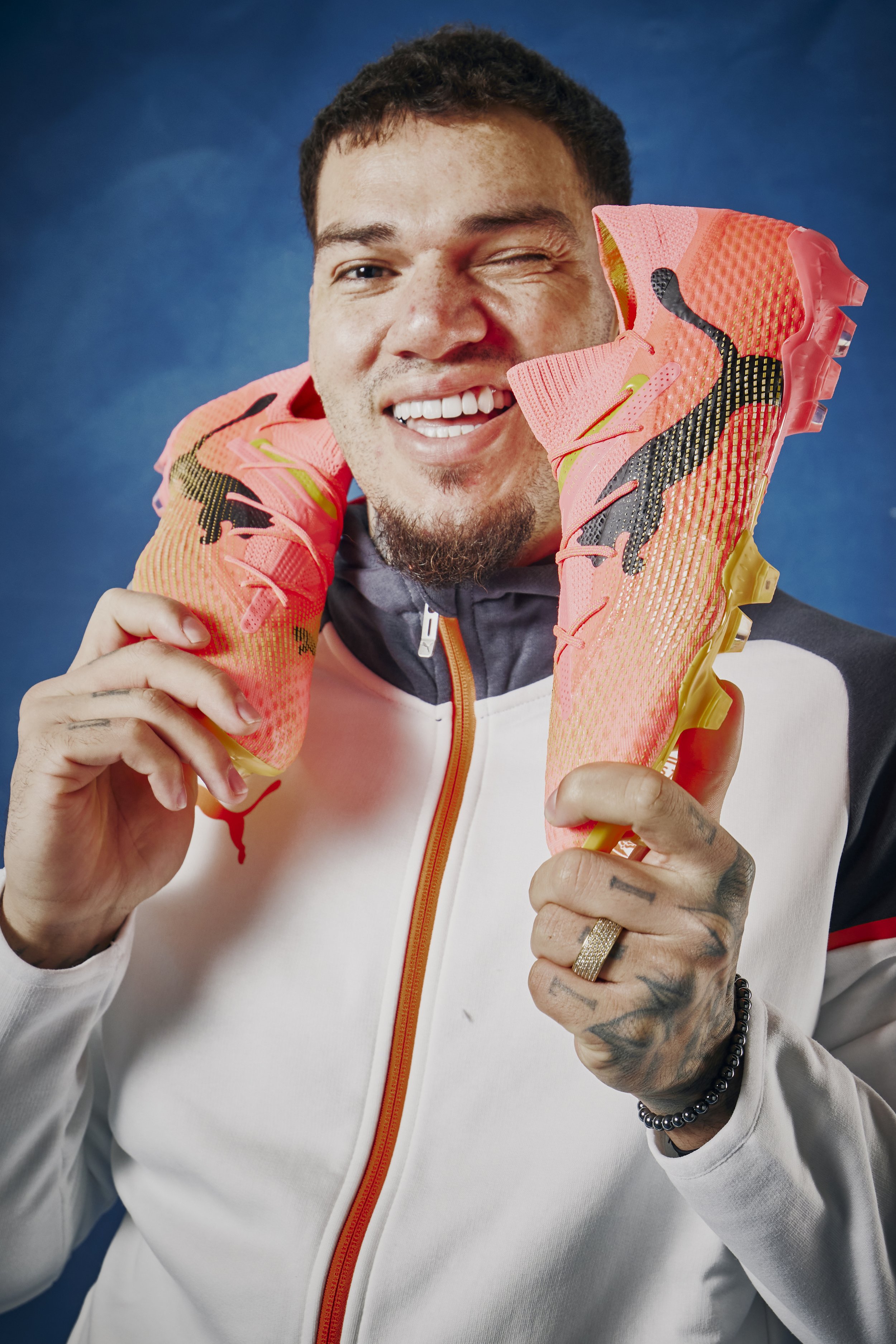 A man winking and smiling while holding a pair of pink running shoes with a black Puma logo and yellow sole near his face. He is wearing a white jacket with a black, gray, and red design and accessories including a ring, beaded bracelet, and tattoos 