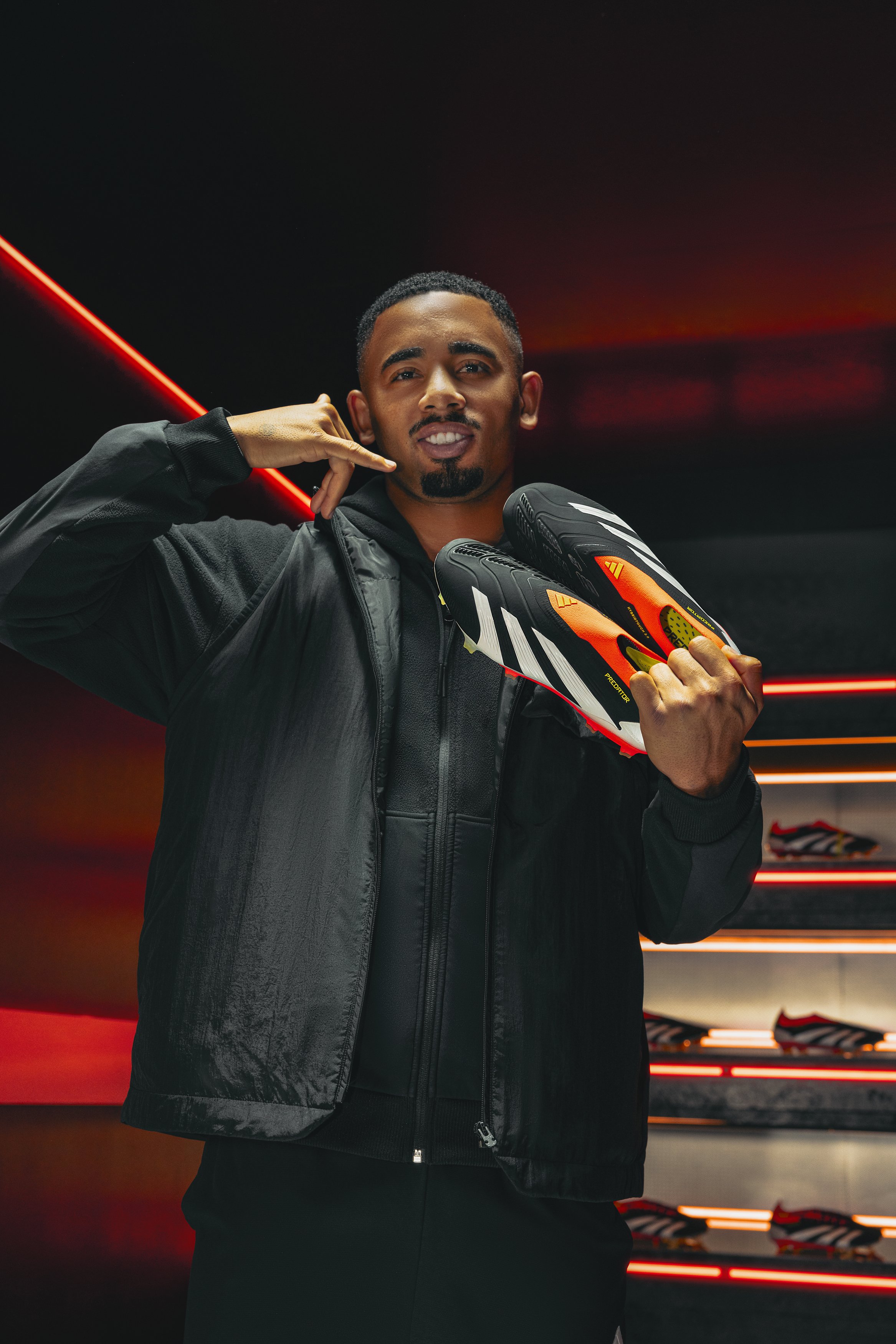 Man in black jacket holding orange and black Nike soccer cleats, standing in front of a red and black background with shelves of similar cleats.