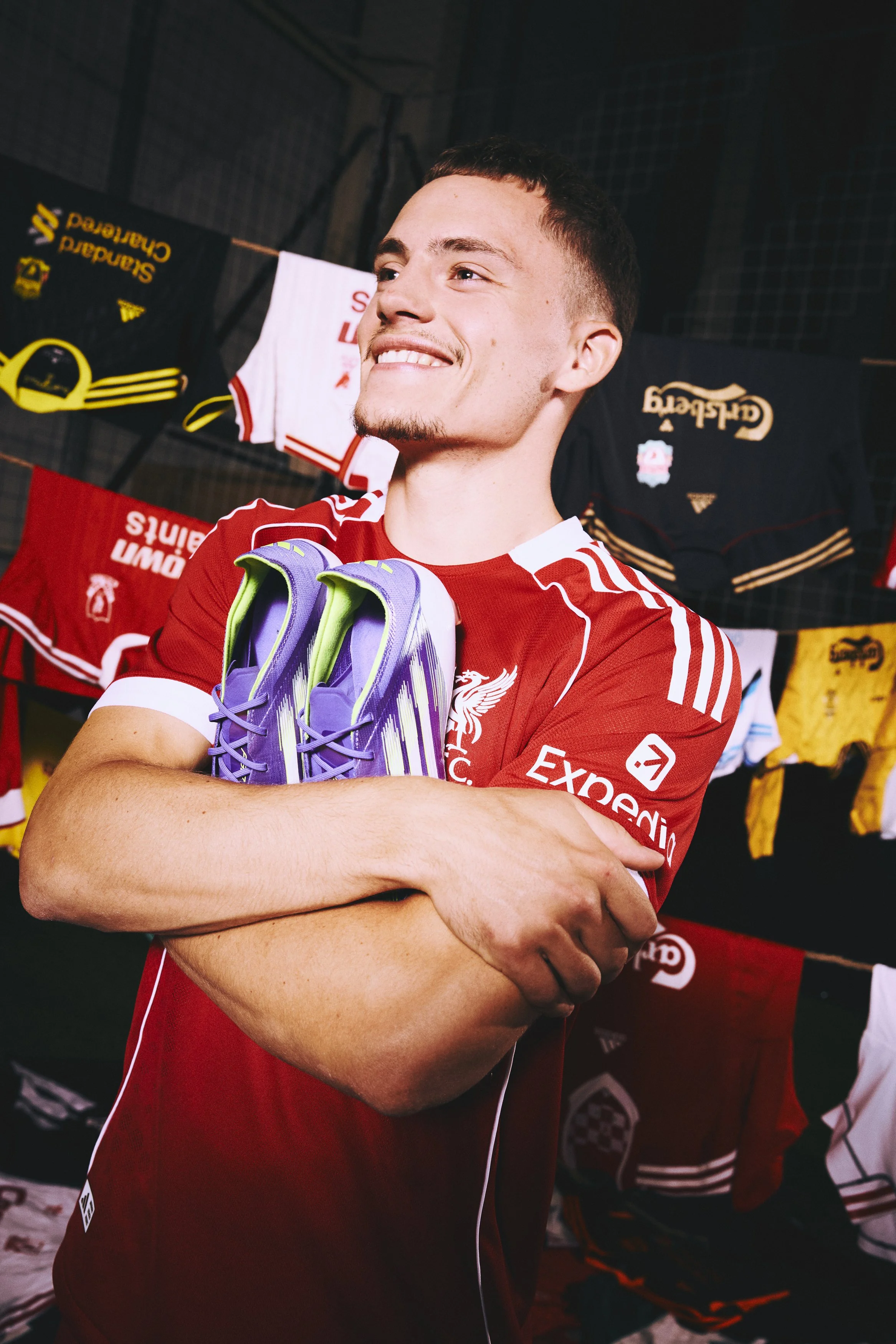 A young man in a red sports jersey is smiling and holding a pair of purple soccer cleats. Behind him, there are various sports jerseys hanging.