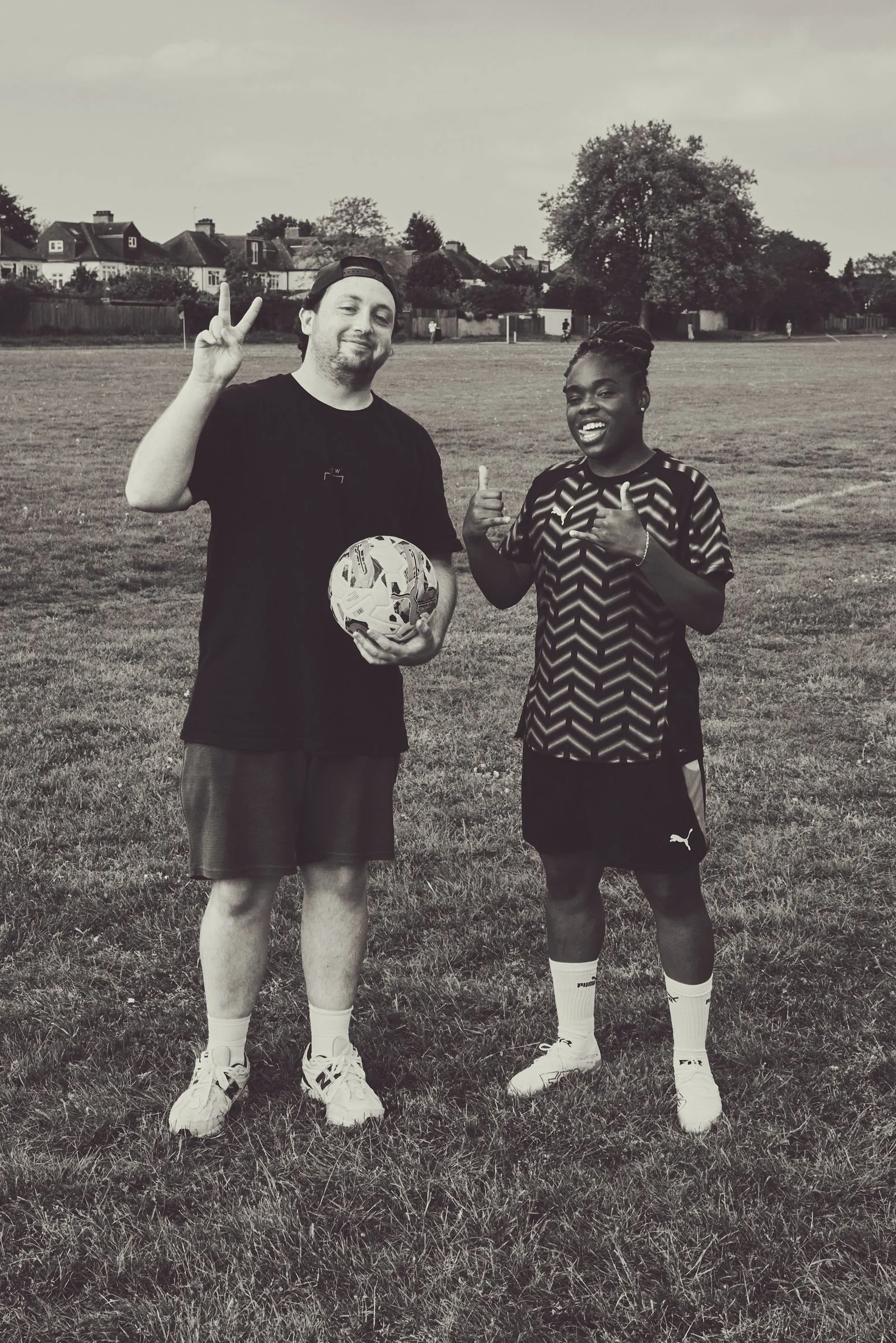 Two men standing on a grassy field, one holding a football, both making peace signs and smiling.