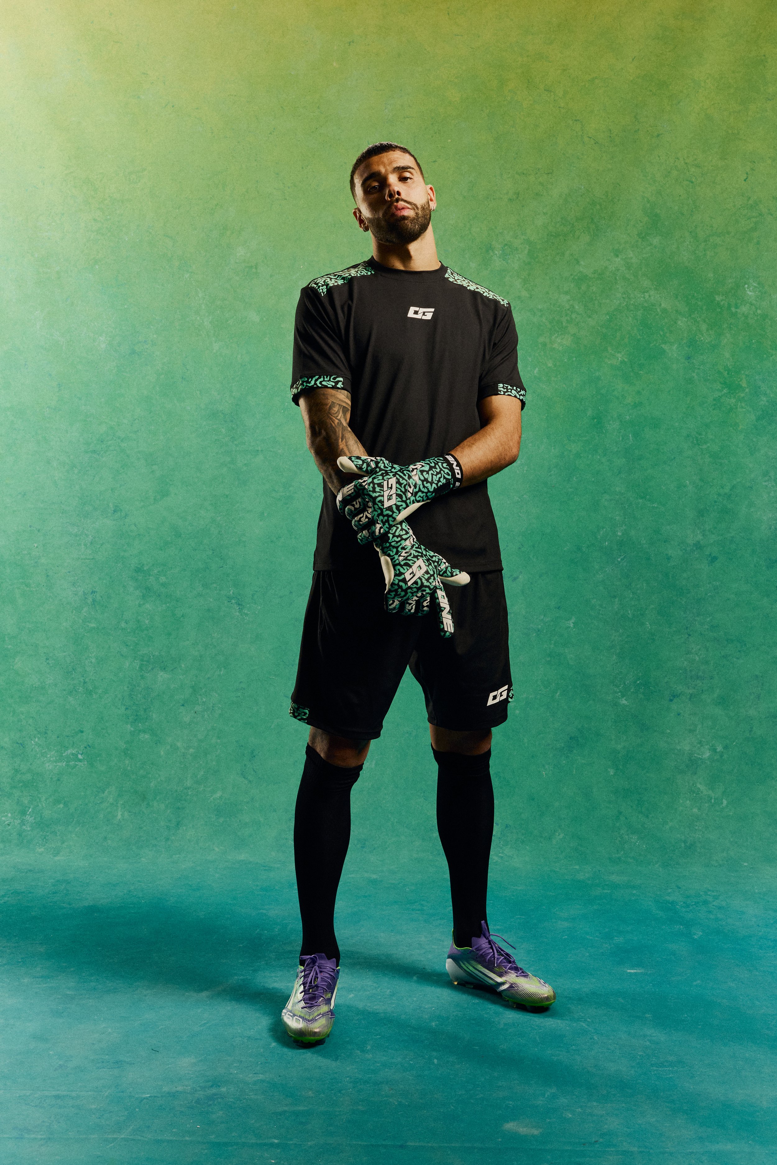 A soccer player stands on a green background, dressed in black sportswear and purple soccer cleats, wearing matching gloves, with a confident expression.