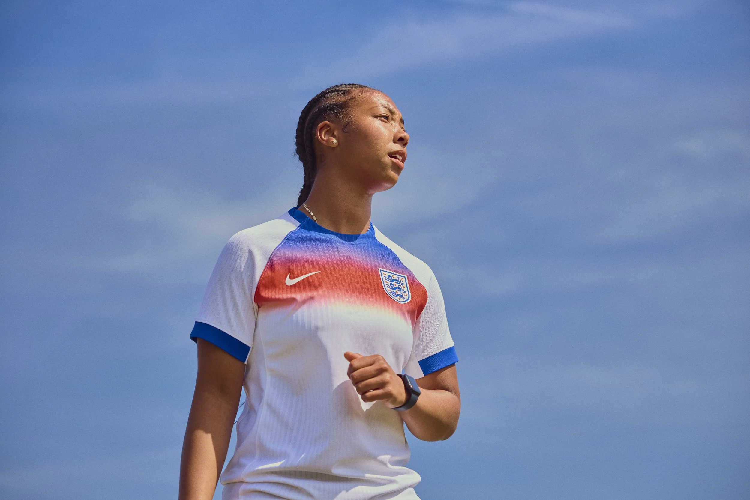 A female soccer player wearing an England national team jersey on the field, with a blue sky in the background.