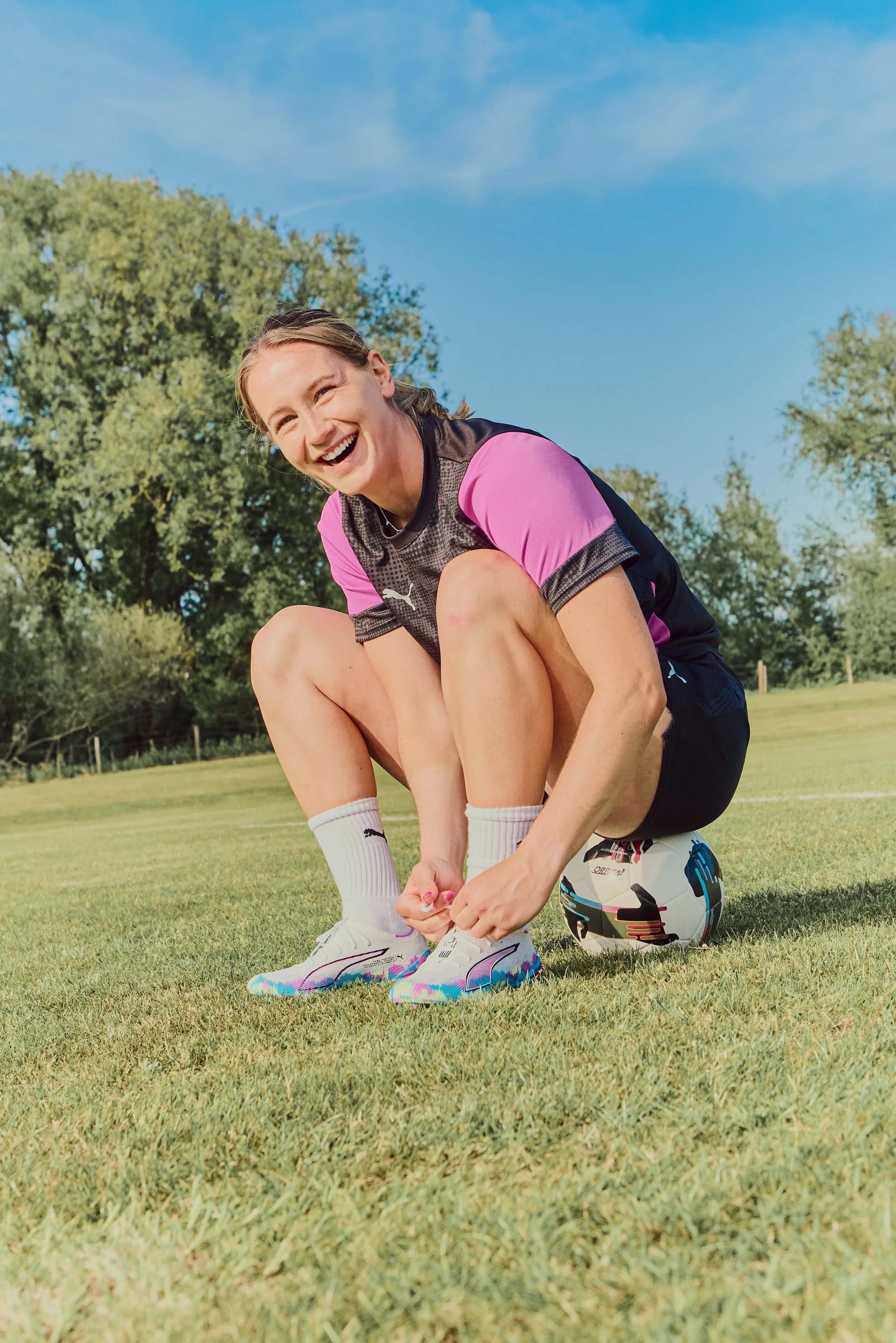 A young woman in athletic attire is smiling and tying her shoelaces on a soccer field, with a soccer ball under her knee and a helmet nearby.