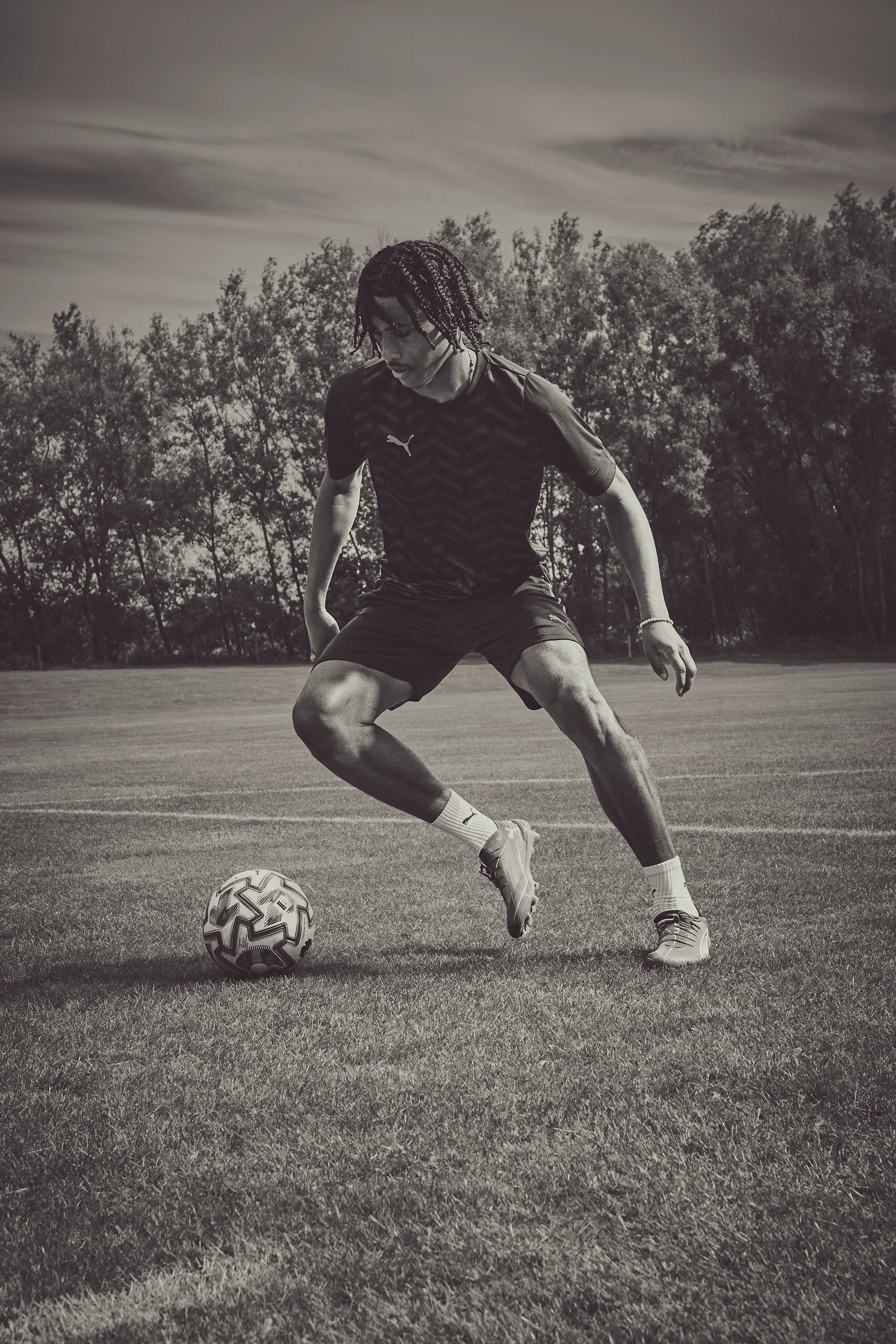 A male soccer player with braided hair wearing a dark sports uniform and soccer cleats, standing on a soccer field with a soccer ball at his feet, outdoors with trees and a cloudy sky in the background.