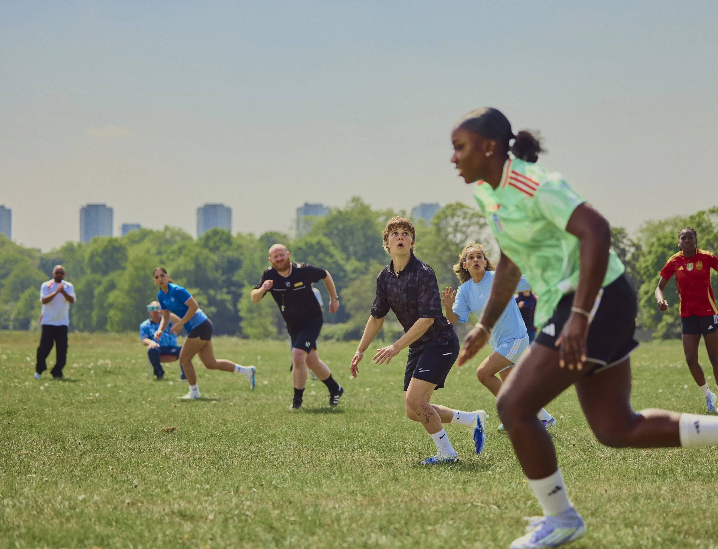 Group of people playing soccer outdoors on a sunny day, with green trees and city buildings in the background.