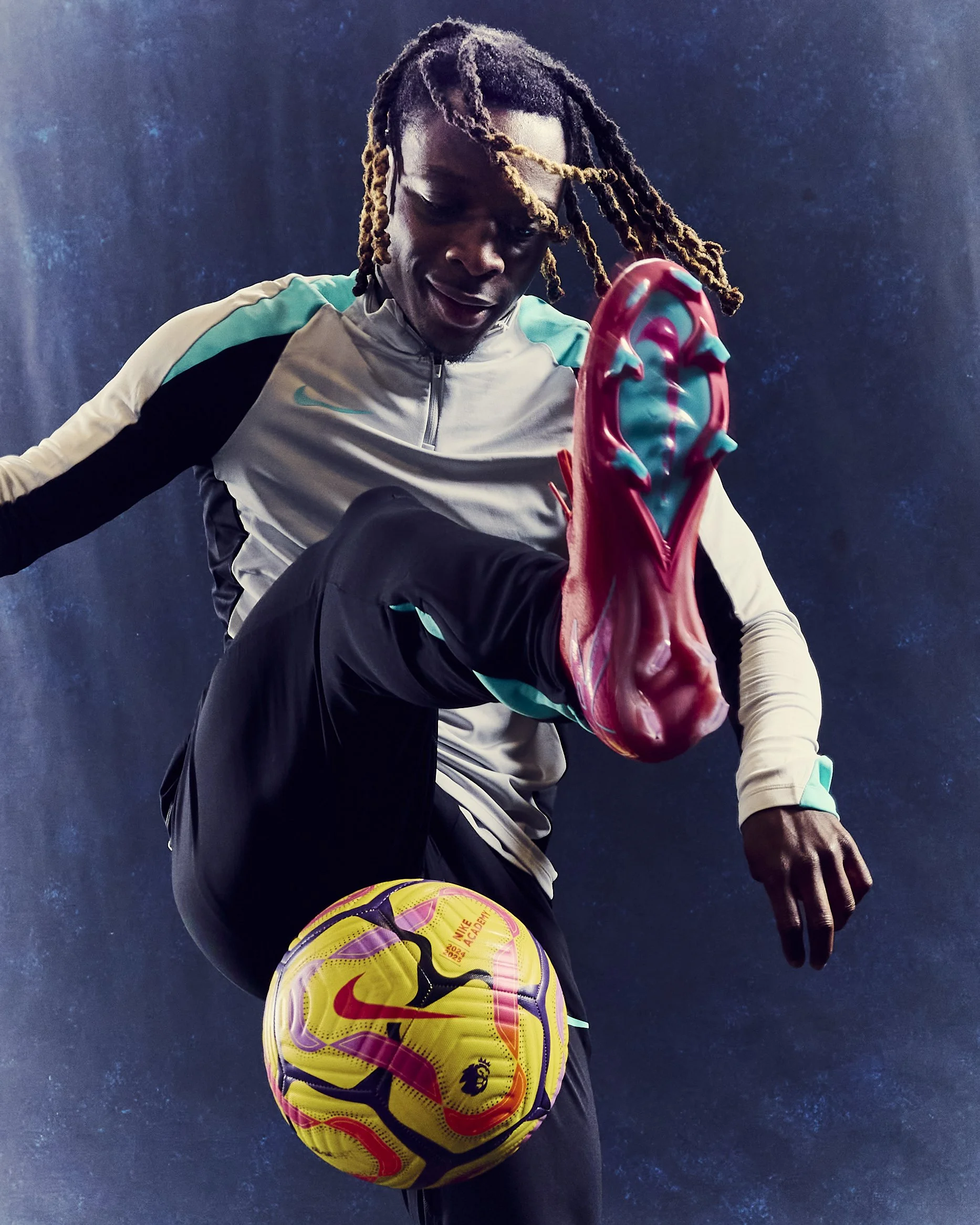 A man with dreadlocks balancing a yellow and purple Nike soccer ball on his knee, wearing a gray and turquoise sports jacket and black pants, against a dark background.