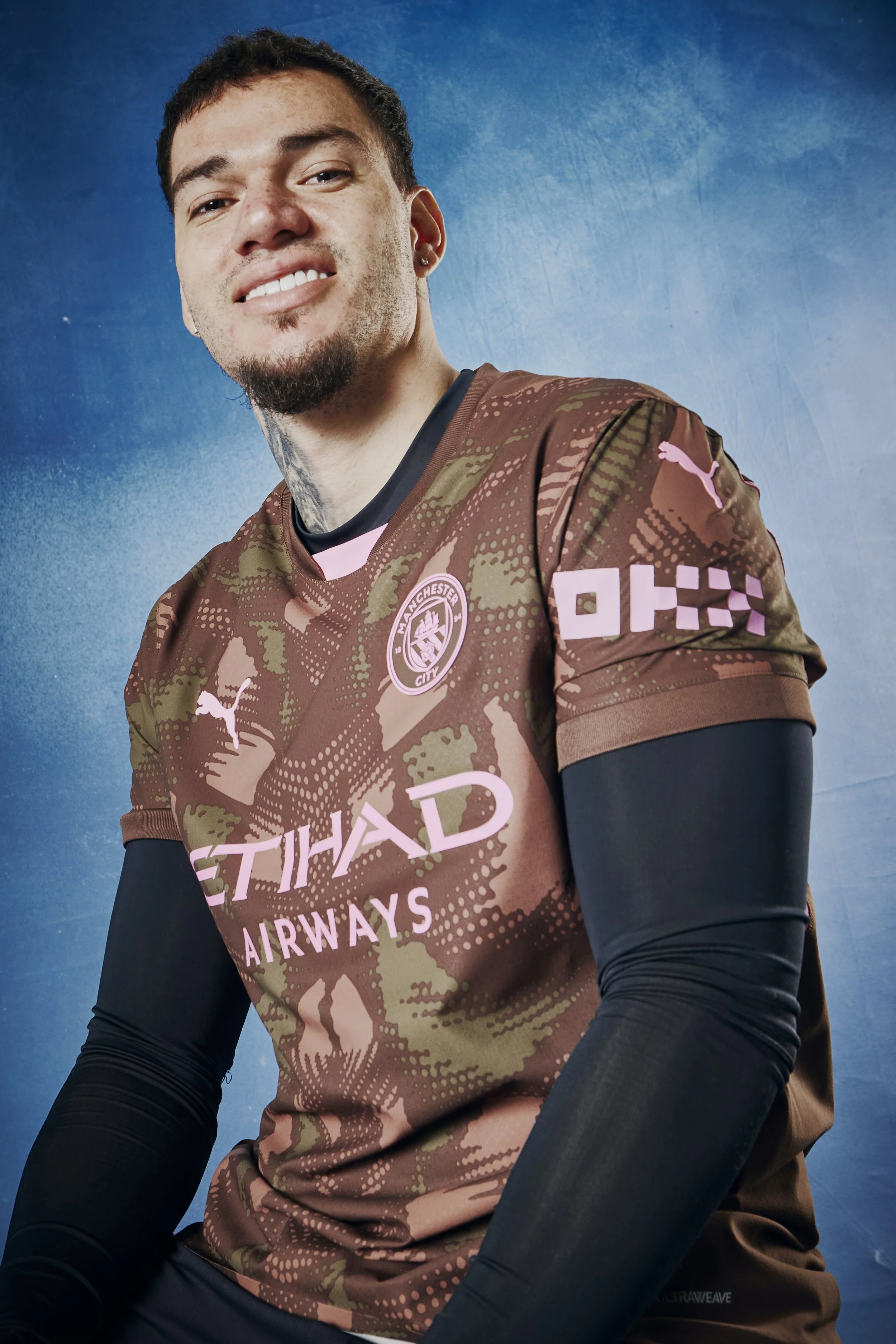 A young man in a Manchester City football jersey with pink branding, posing against a blue background, smiling at the camera.