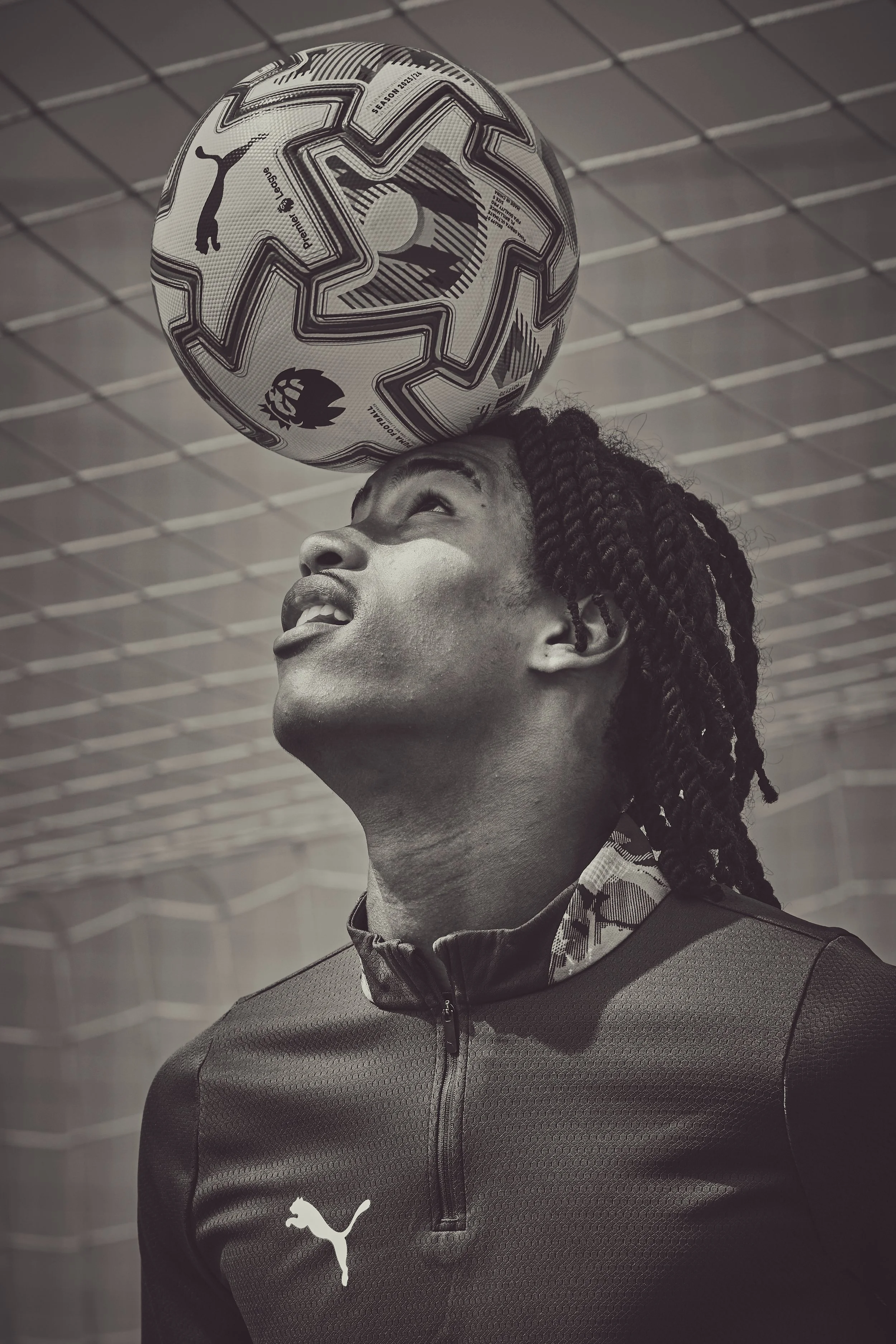 A young man with dreadlocks balancing a soccer ball on his forehead indoors, wearing a sports jacket with a Puma logo.