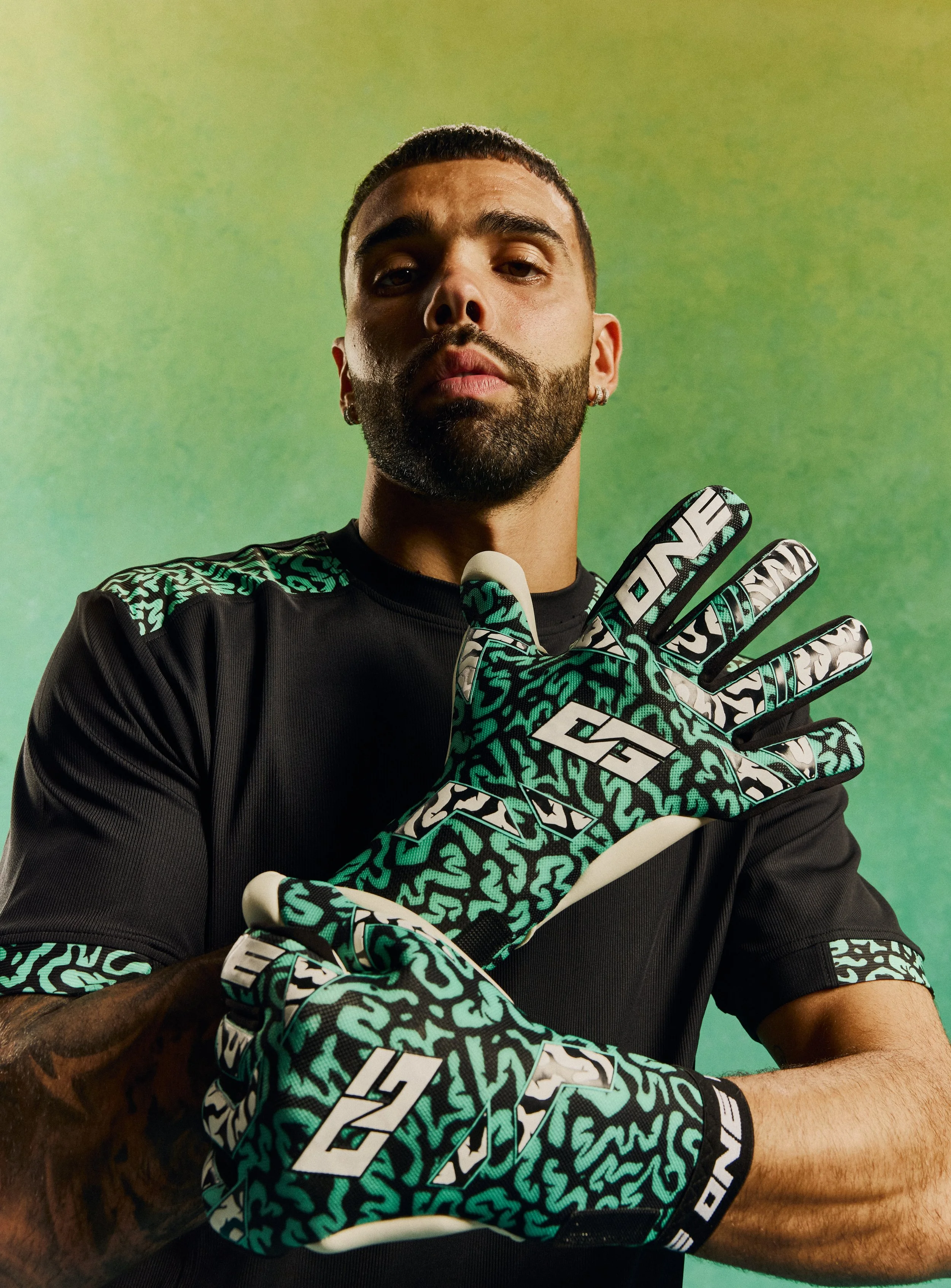 A man wearing black sports attire and patterned gloves with teal and white designs, featuring the brand name "ONE". He has a beard, short hair, and is posing with a serious expression against a green gradient background.