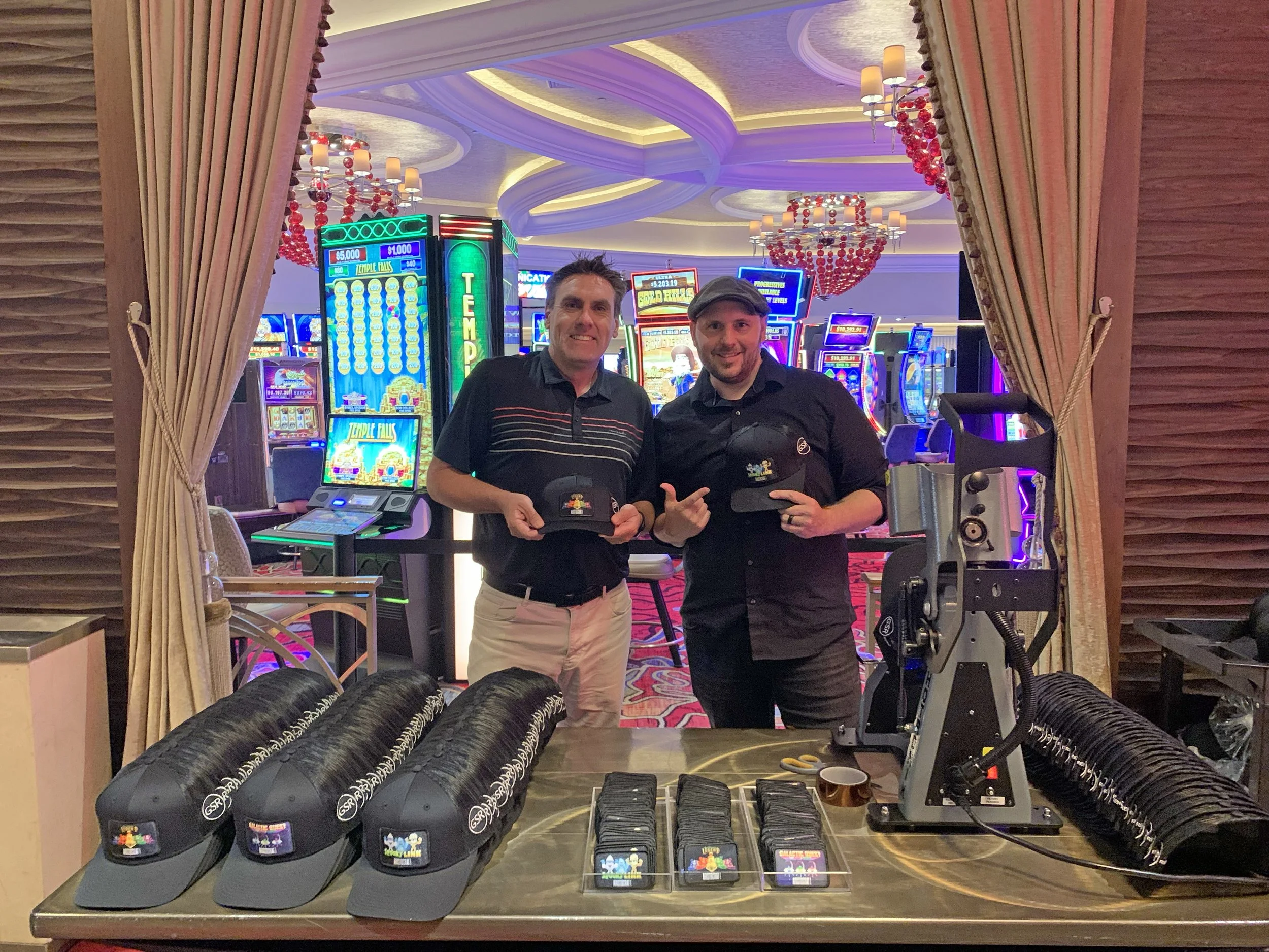 Two men standing behind a table with hats, patches, and equipment in a casino with slot machines in the background.