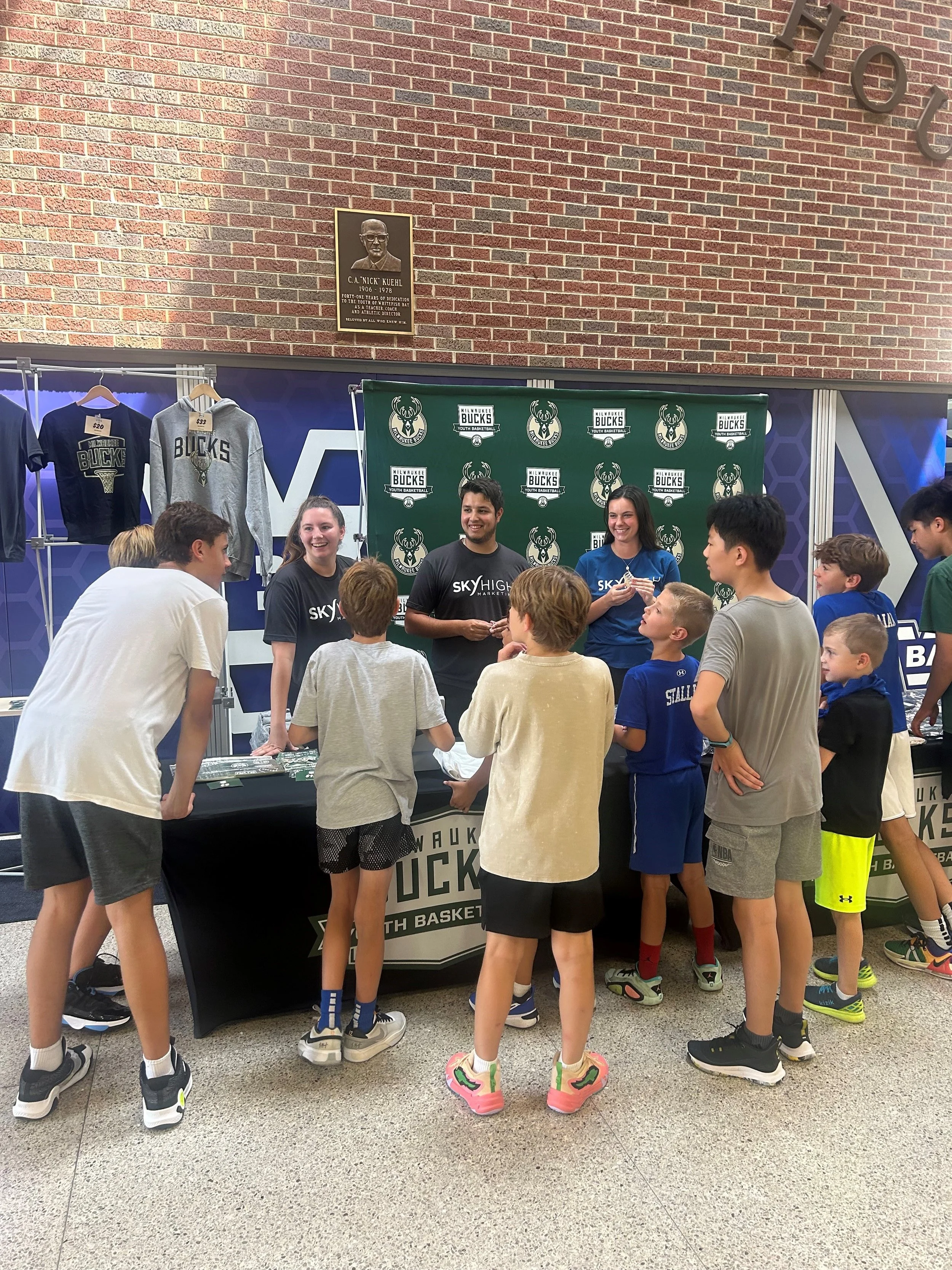 Children gathered around a basketball autograph signing booth with Milwaukee Bucks branding, where three adults are standing and talking to the kids.