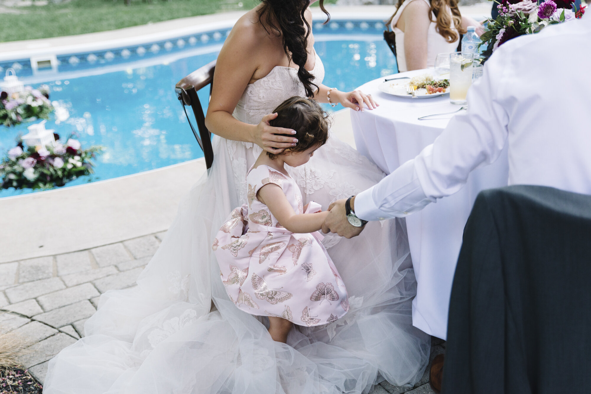 Candid photo of a child playing on a bride's wedding dress during a backyard reception as the bride embraces the moment unbothered